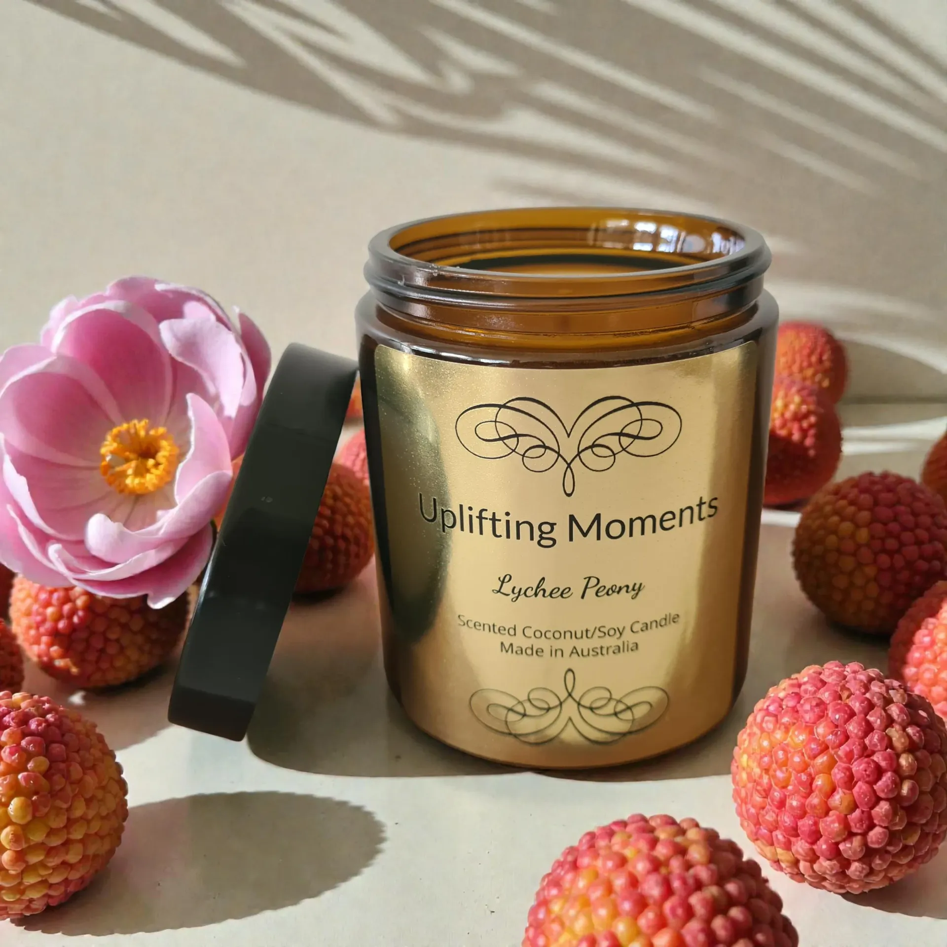 Gold candle jar labeled “Uplifting Moments” with pink flower petals and blossoms on a wooden table.