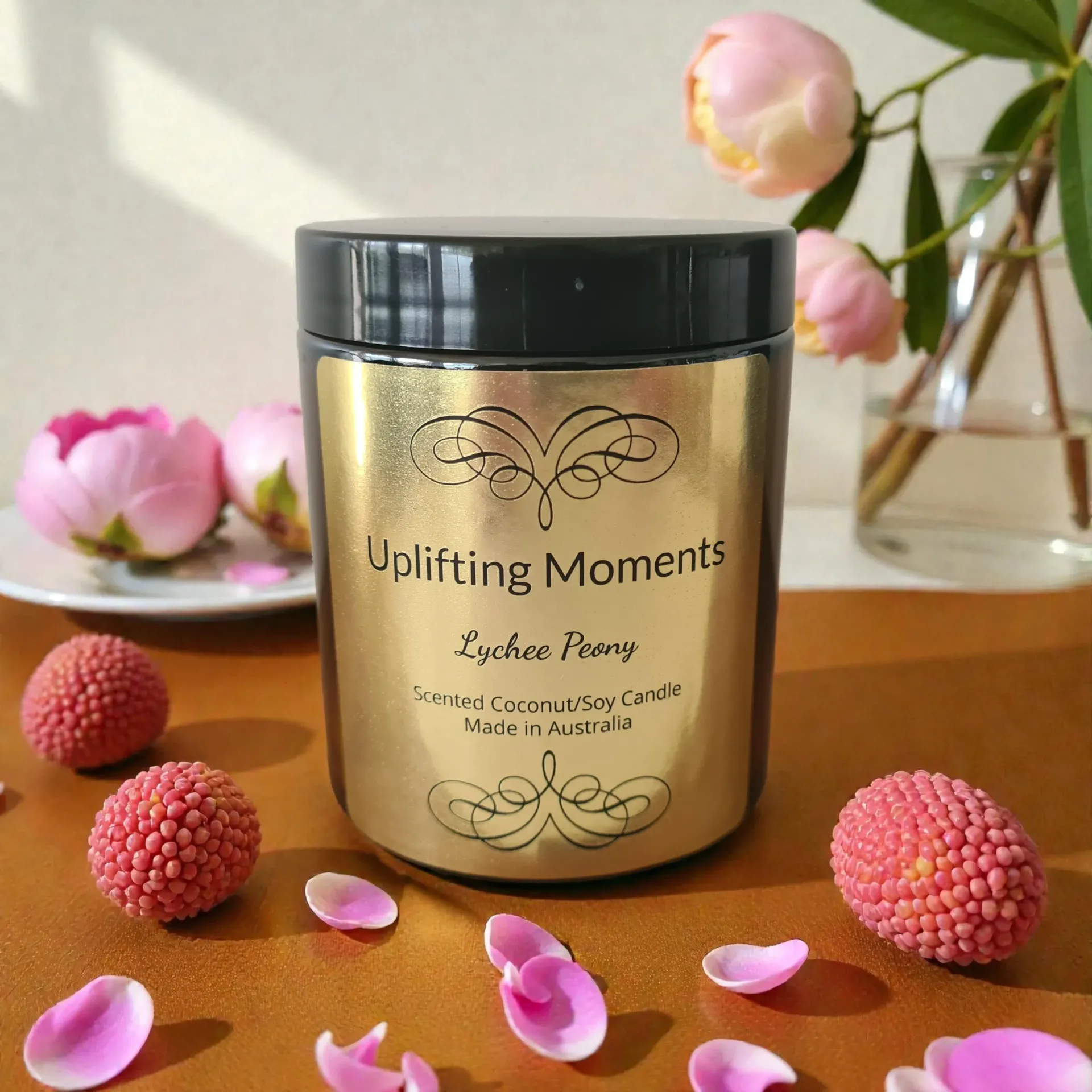 Gold candle jar labeled “Uplifting Moments” with pink flower petals and blossoms on a wooden table.