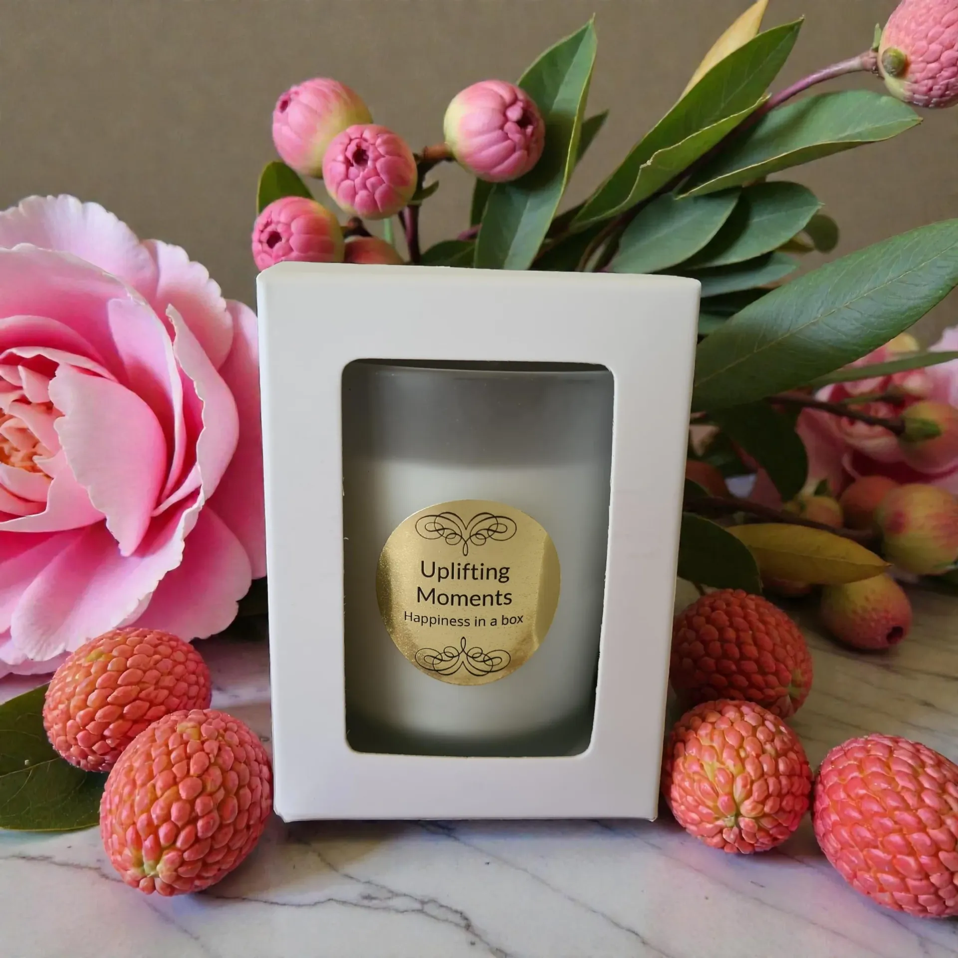 Frosted candle labeled “Uplifting Moments” surrounded by pink lychees and a pink flower on a wooden surface