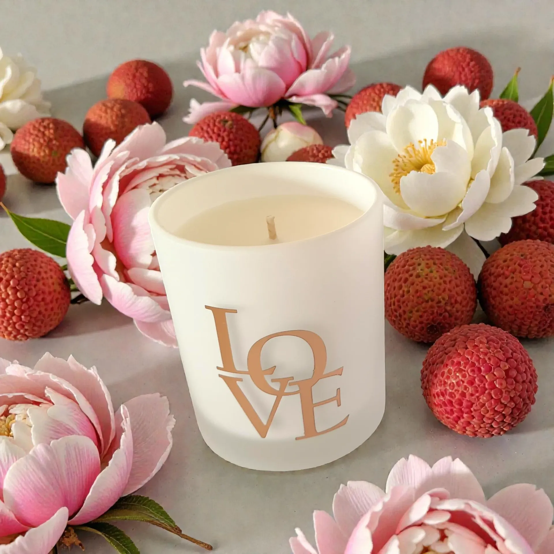 White candle with rose gold “LOVE” text surrounded by pink flowers and red berries