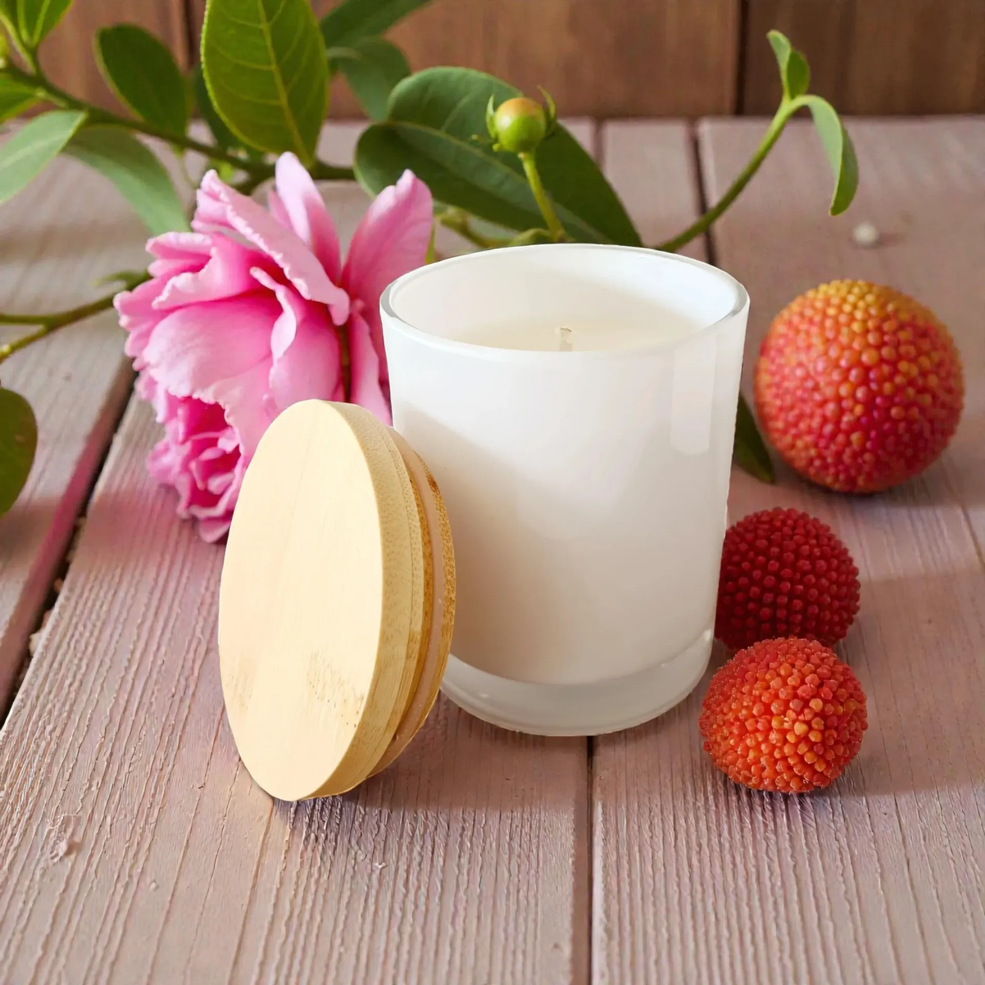 White candle with pink flower, round wooden lid, and berry-like fruits on a wooden surface