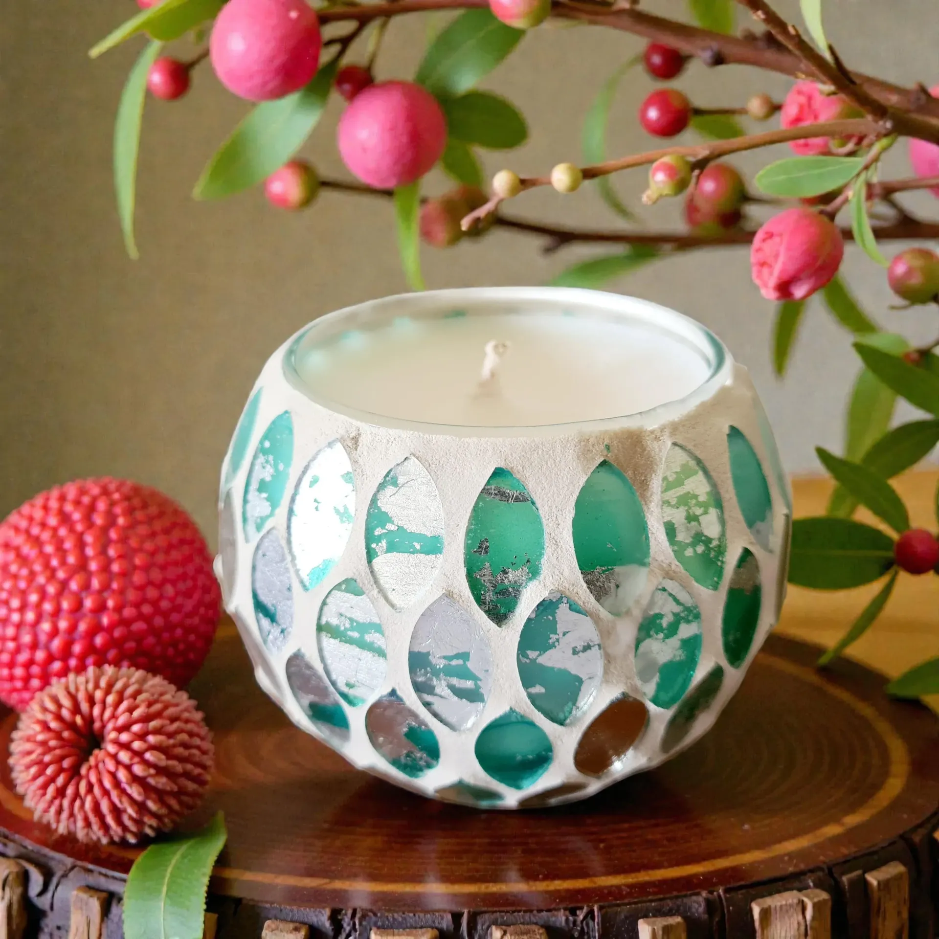 White candle in a turquoise mosaic holder on a wooden slice, with pink flowers in the background