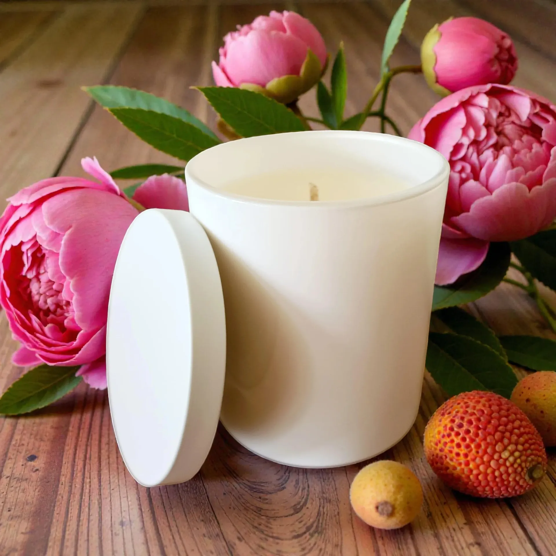White candle jar with lid beside pink flowers and fruit on a wooden table