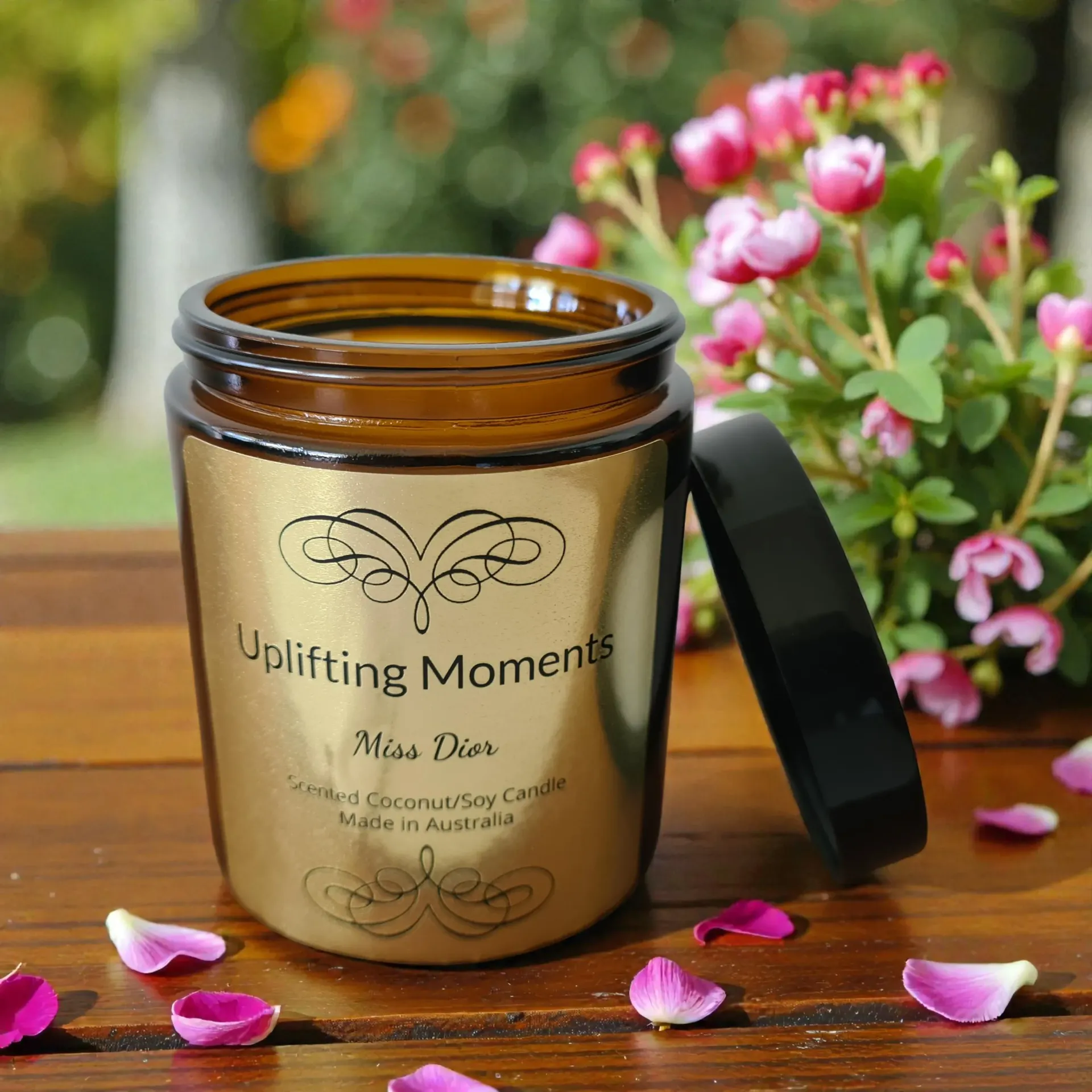 Gold candle jar with black lid labeled “Uplifting Moments,” surrounded by pink flowers and petals on a wooden surface