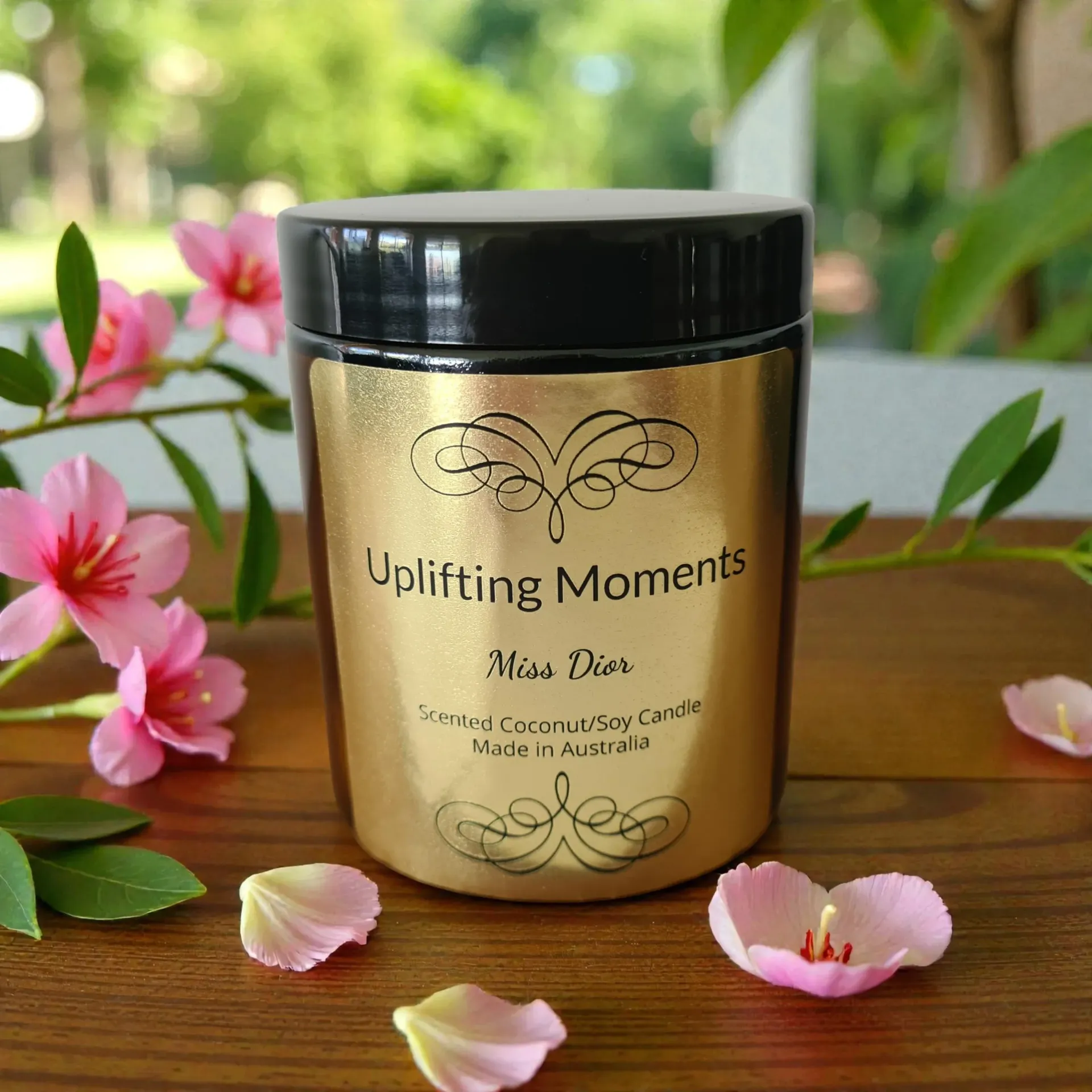 Gold candle jar with black lid labeled “Uplifting Moments,” surrounded by pink flowers and petals on a wooden surface