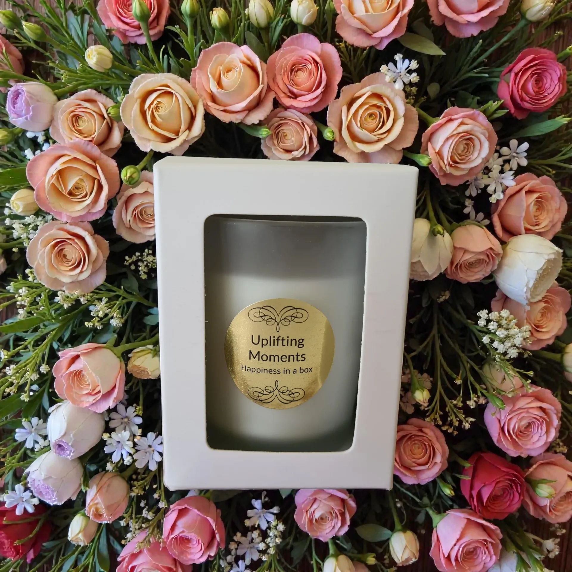 Frosted candle labeled “Uplifting Moments” surrounded by pink and red roses on a wooden surface