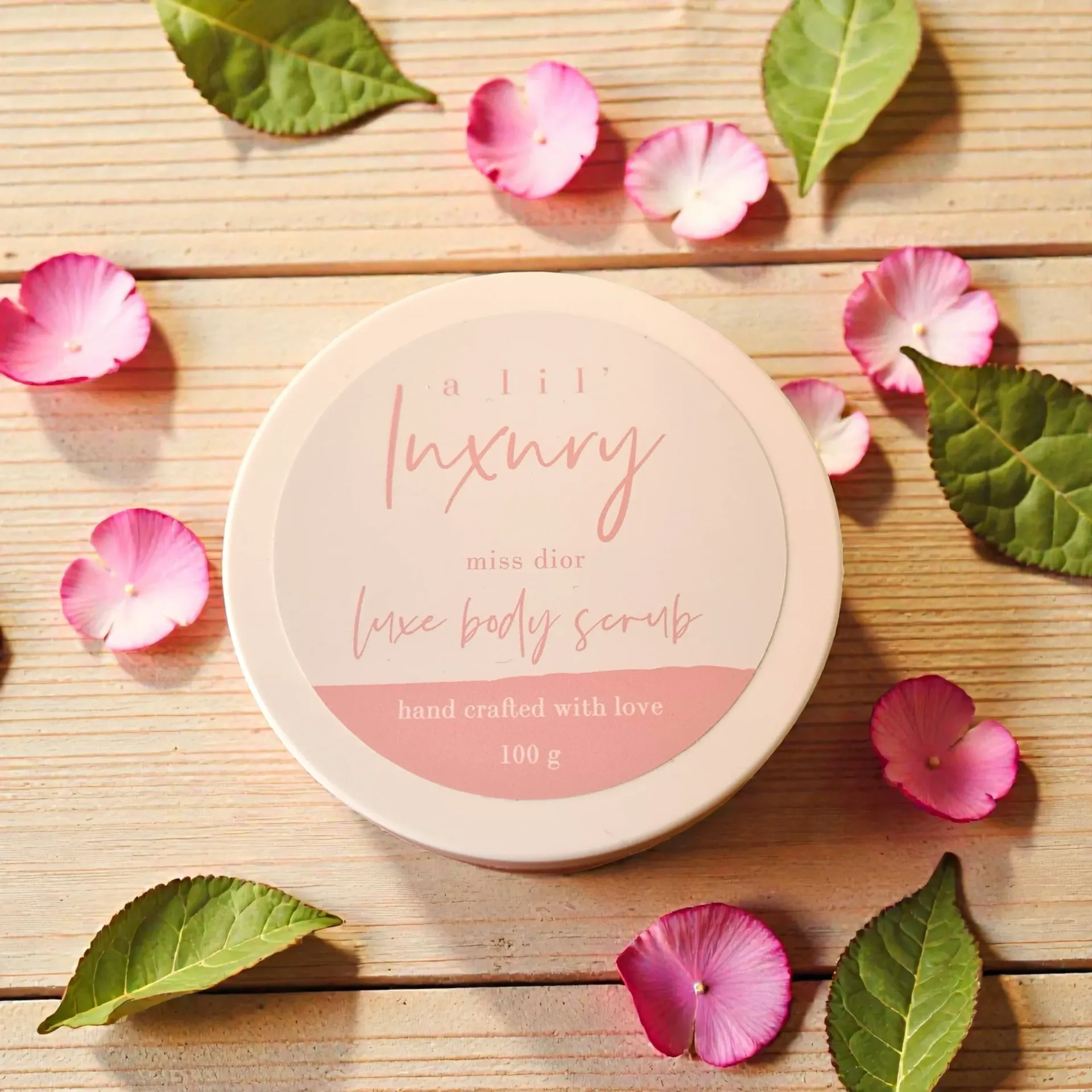 Pink round scrub jar with floral label, surrounded by scattered rose petals on a wooden surface.