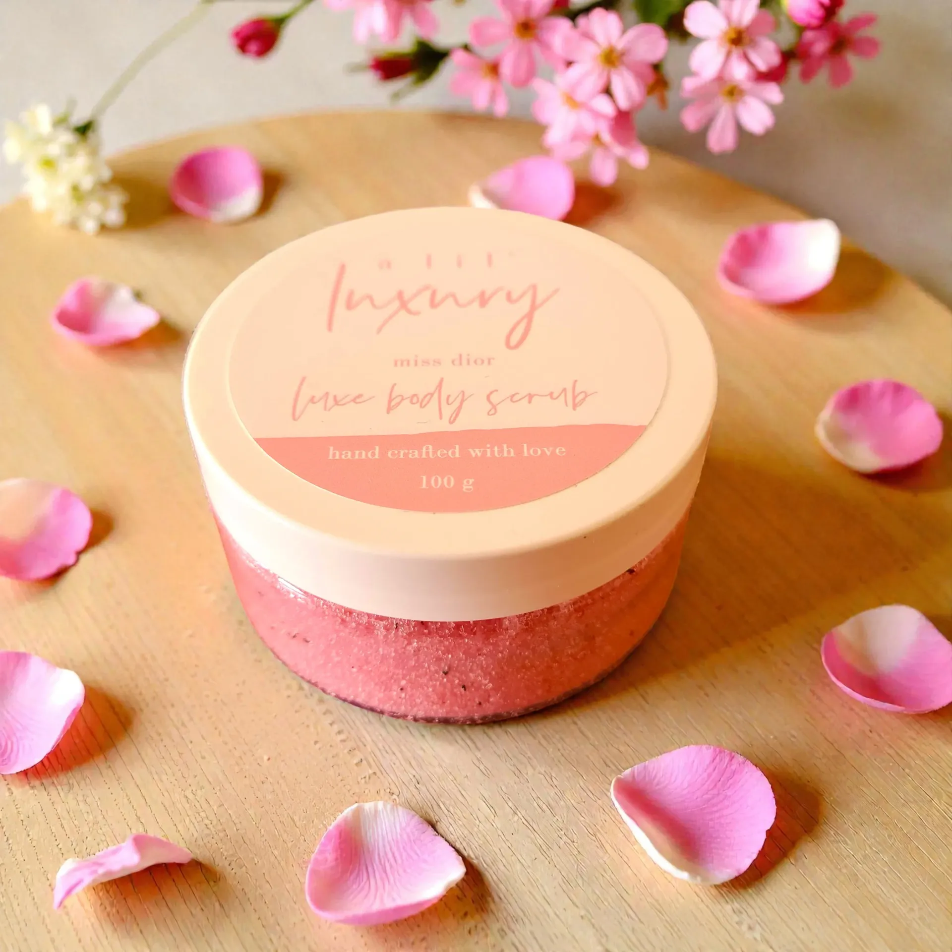 Pink round scrub jar with floral label, surrounded by scattered rose petals on a wooden surface.