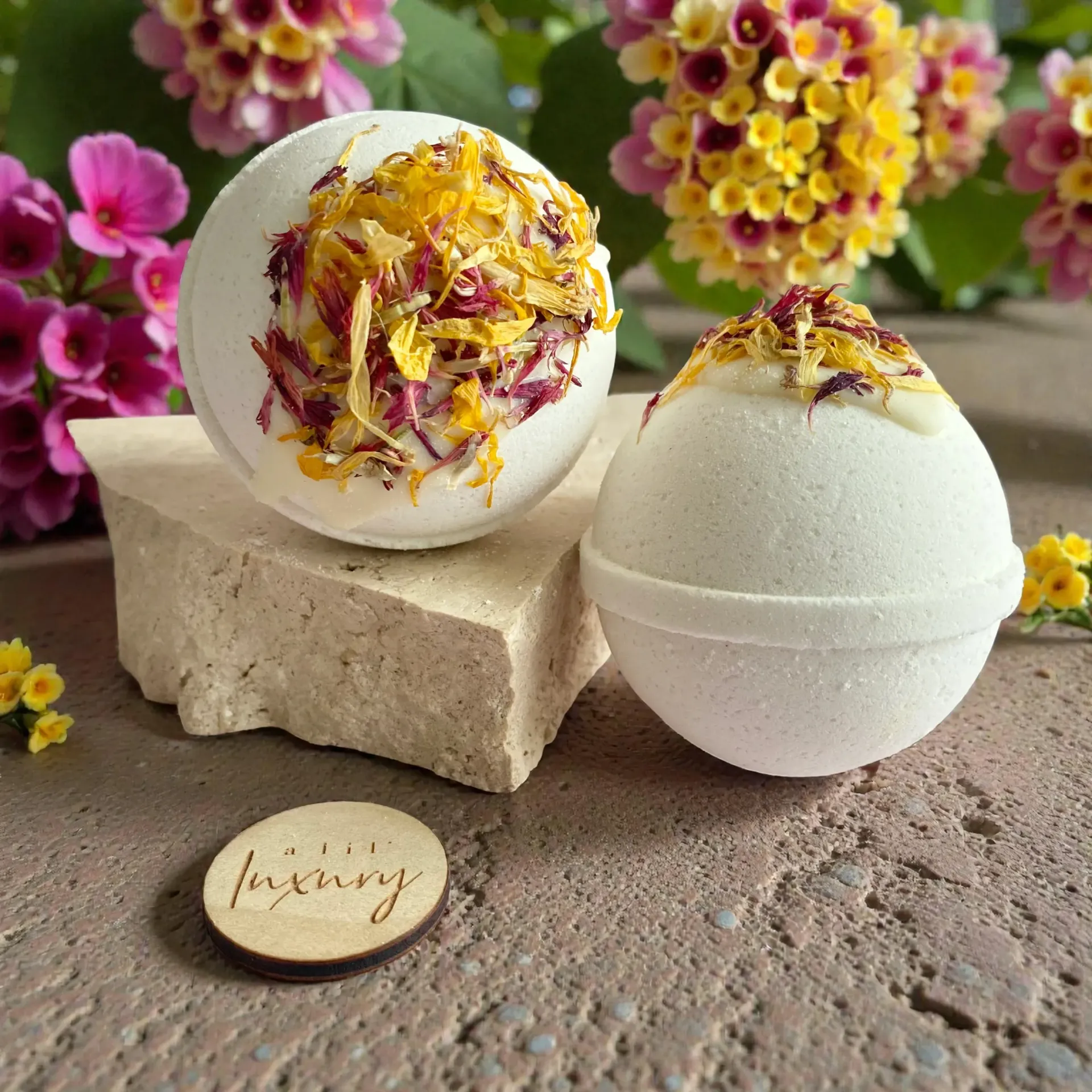 Two white bath bombs with dried flowers on a stone block, surrounded by small blooms.