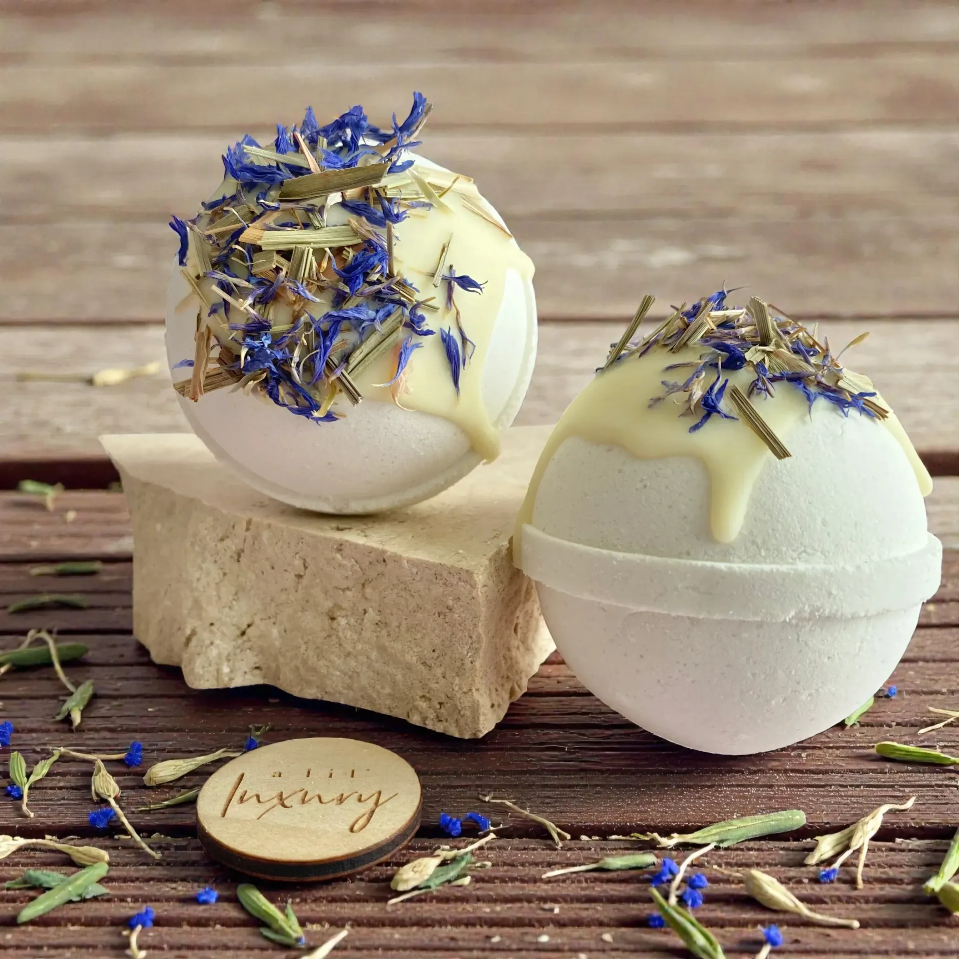 Two white bath bombs topped with yellow drizzle and purple flowers on a wooden surface