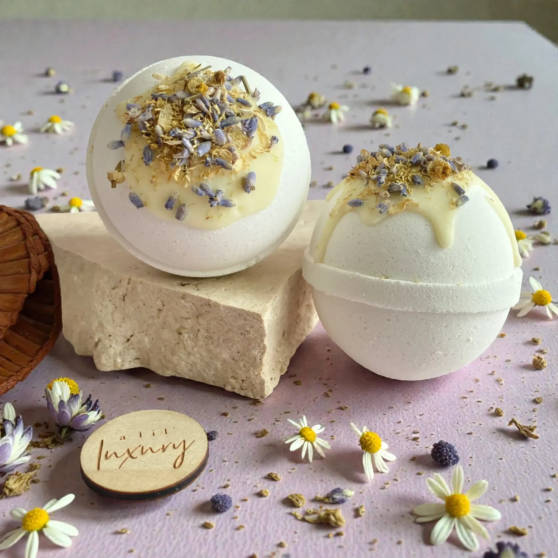 Two white bath bombs with dried flowers on a beige block, surrounded by scattered daisies and a lavender soap bar