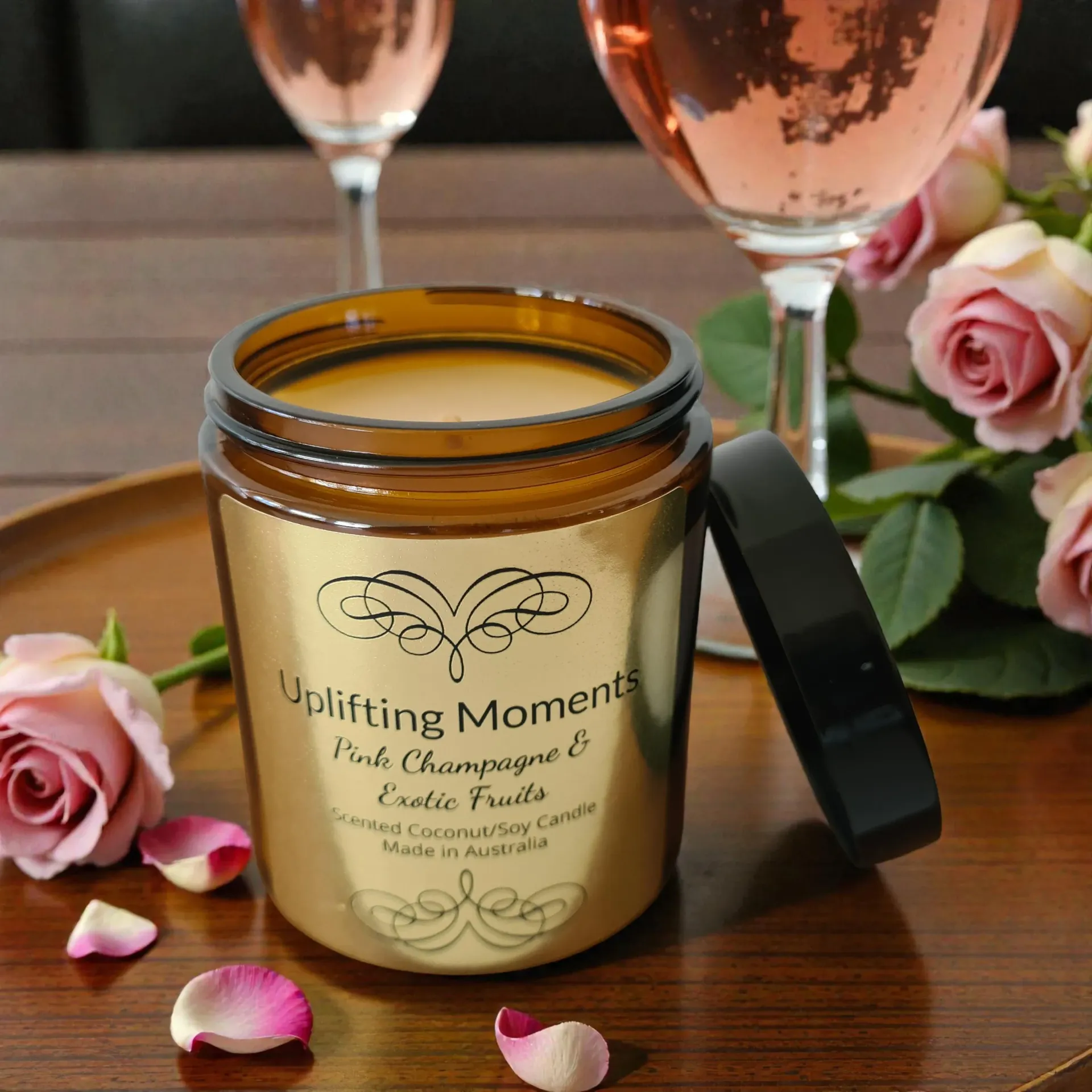 A candle jar with black lid, pink roses, scattered petals, and a champagne flute on a wooden surface.