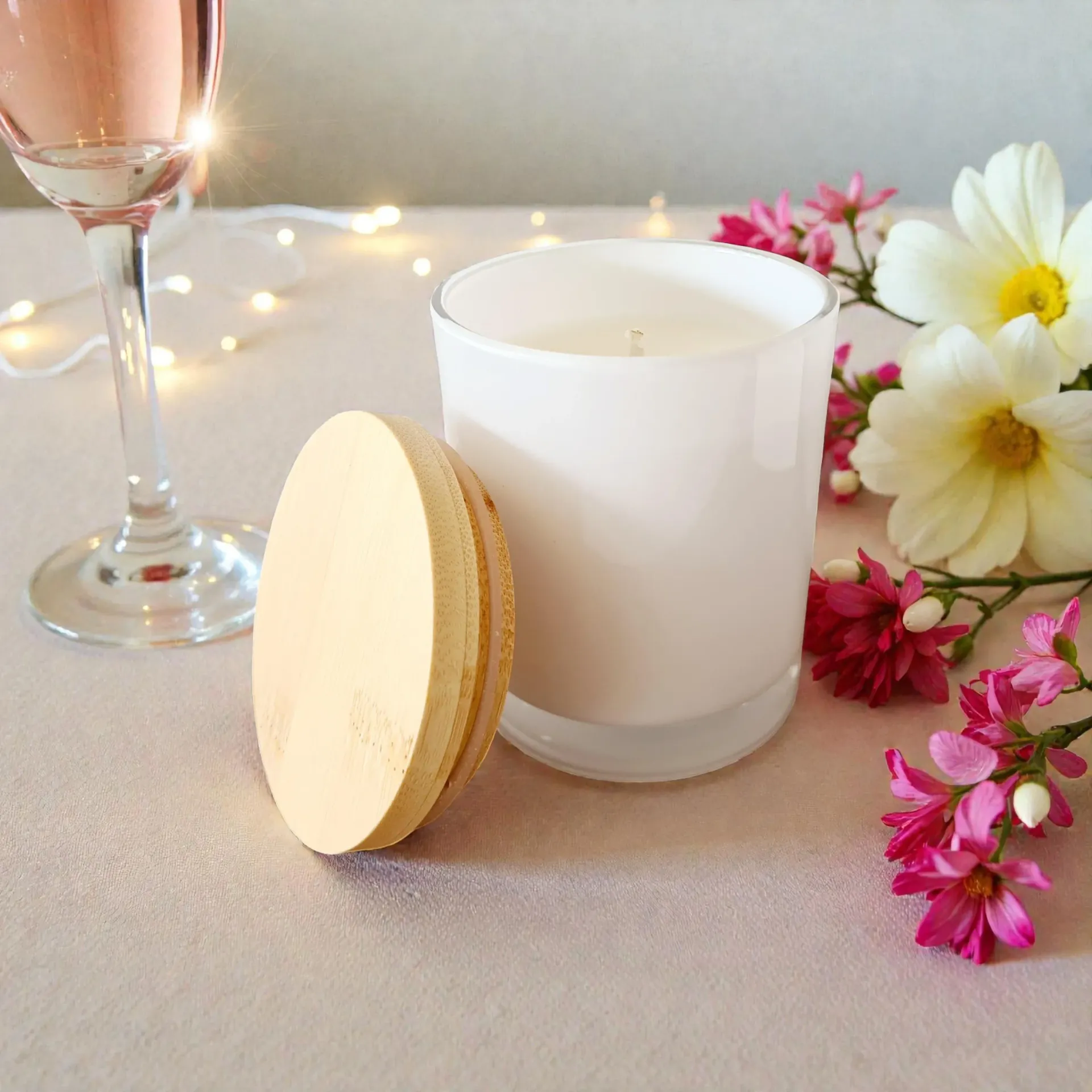 White candle with wooden lid, wine glass, and pink flowers on a soft beige surface