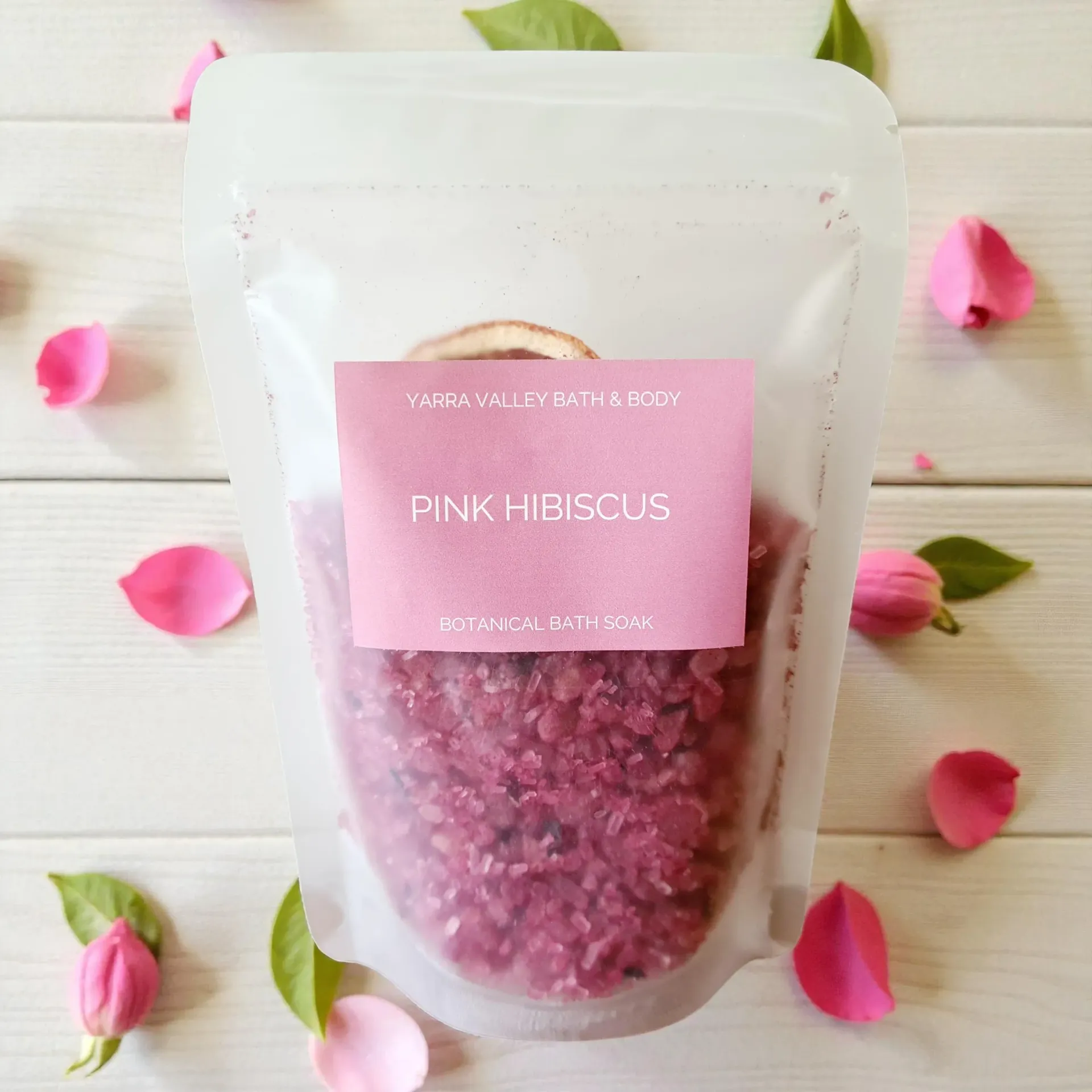 Pink hibiscus bath salts in a clear pouch with pink label, surrounded by pink petals on a white background
