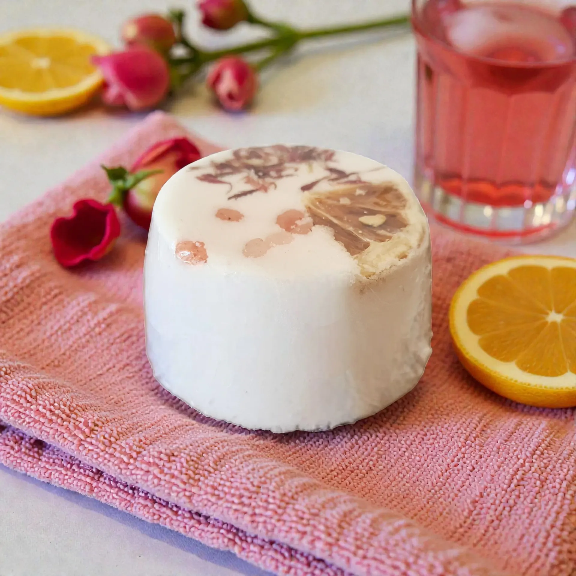 White bath bomb on pink towel with orange slices, red drink, and flowers on a pastel table