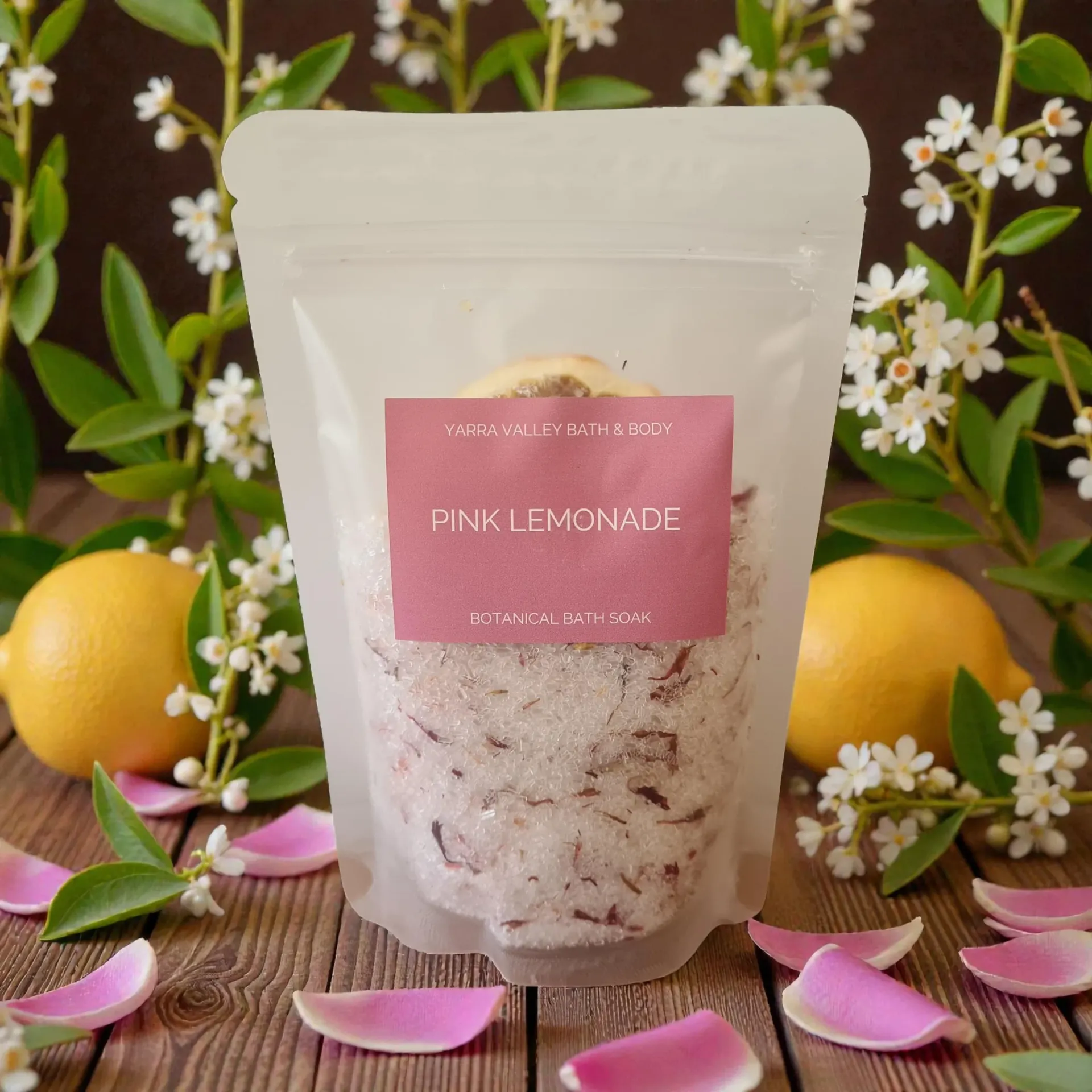 Pink Lemonade bath soak pouch with pink label, surrounded by lemons, flowers, and pink petals