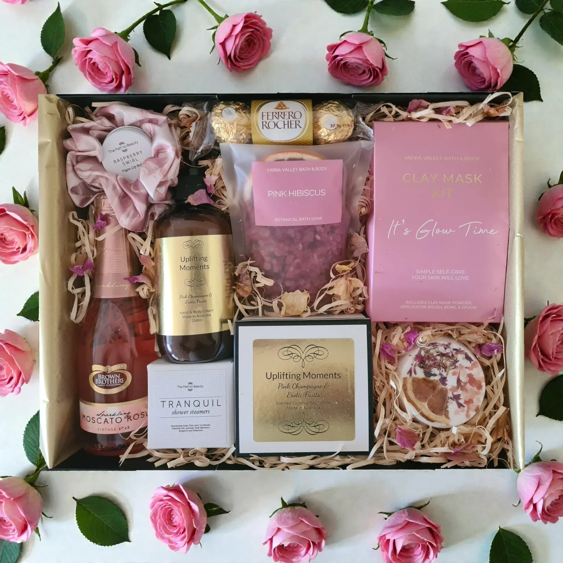 Gift box with pink roses, candles, body products, treats, and a champagne bottle on a white background