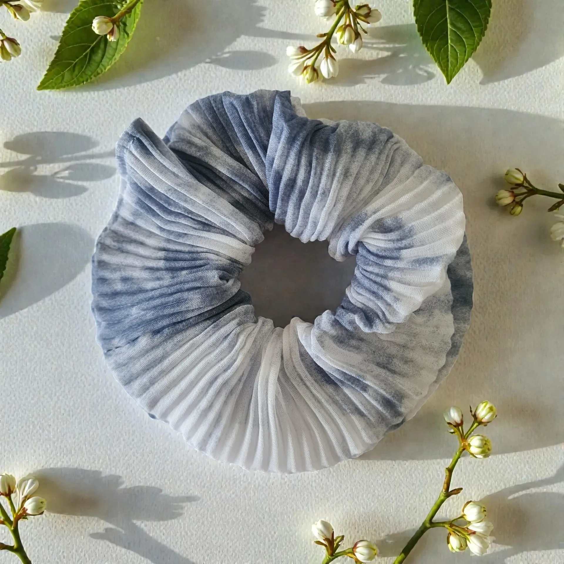 Blue and white pleated fabric scrunchie on a light surface with scattered green leaves and white flowers