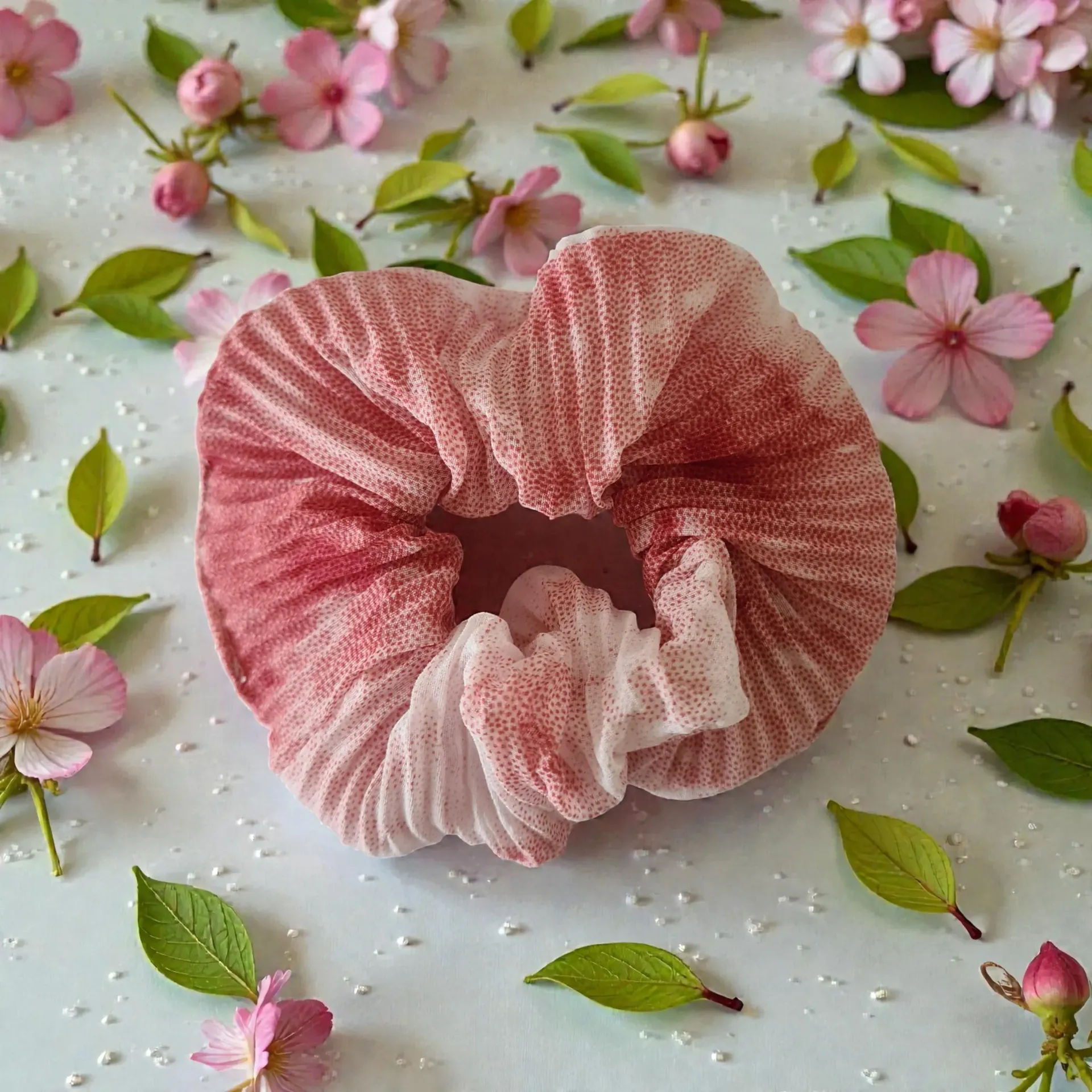 Pink scrunchie on a white textured surface scattered with small pink flowers and green leaves