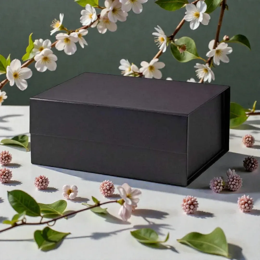 Black rectangular gift box surrounded by white blossoms and green leaves on a gray surface
