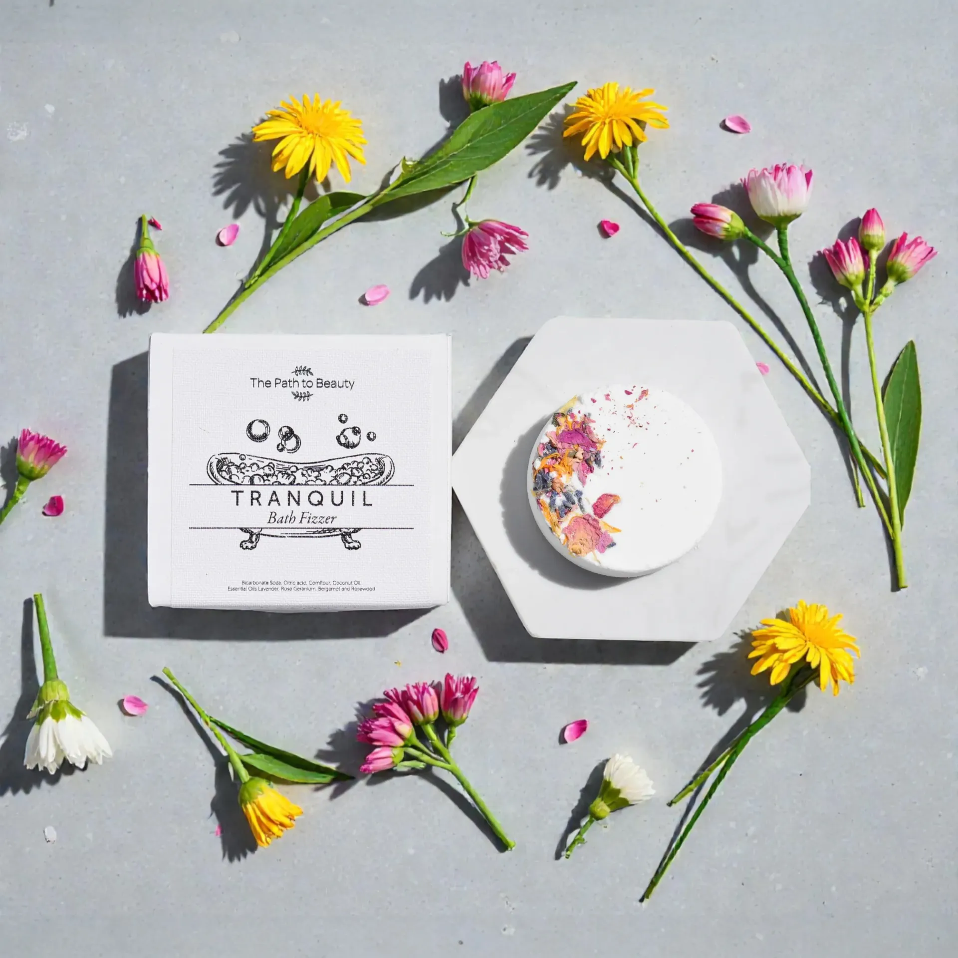 Flat-lay of wildflowers surrounding a white bath fizzer with petals and round box on a gray background