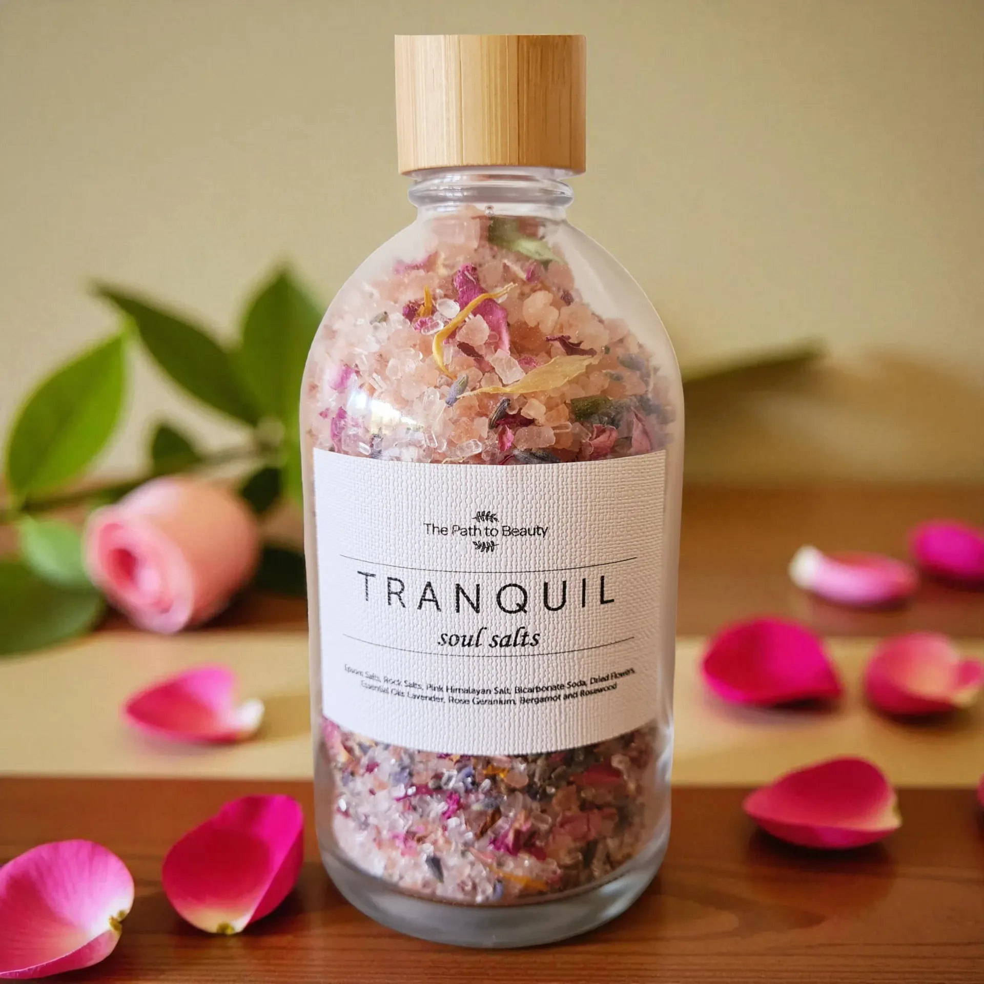 Glass bottle of pink bath salts labeled “Tranquil,” with rose petals on a wooden table