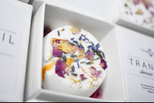 A round, white shower steamer topped with dried rose petals and lavender buds, nestled in a white cardboard gift box.