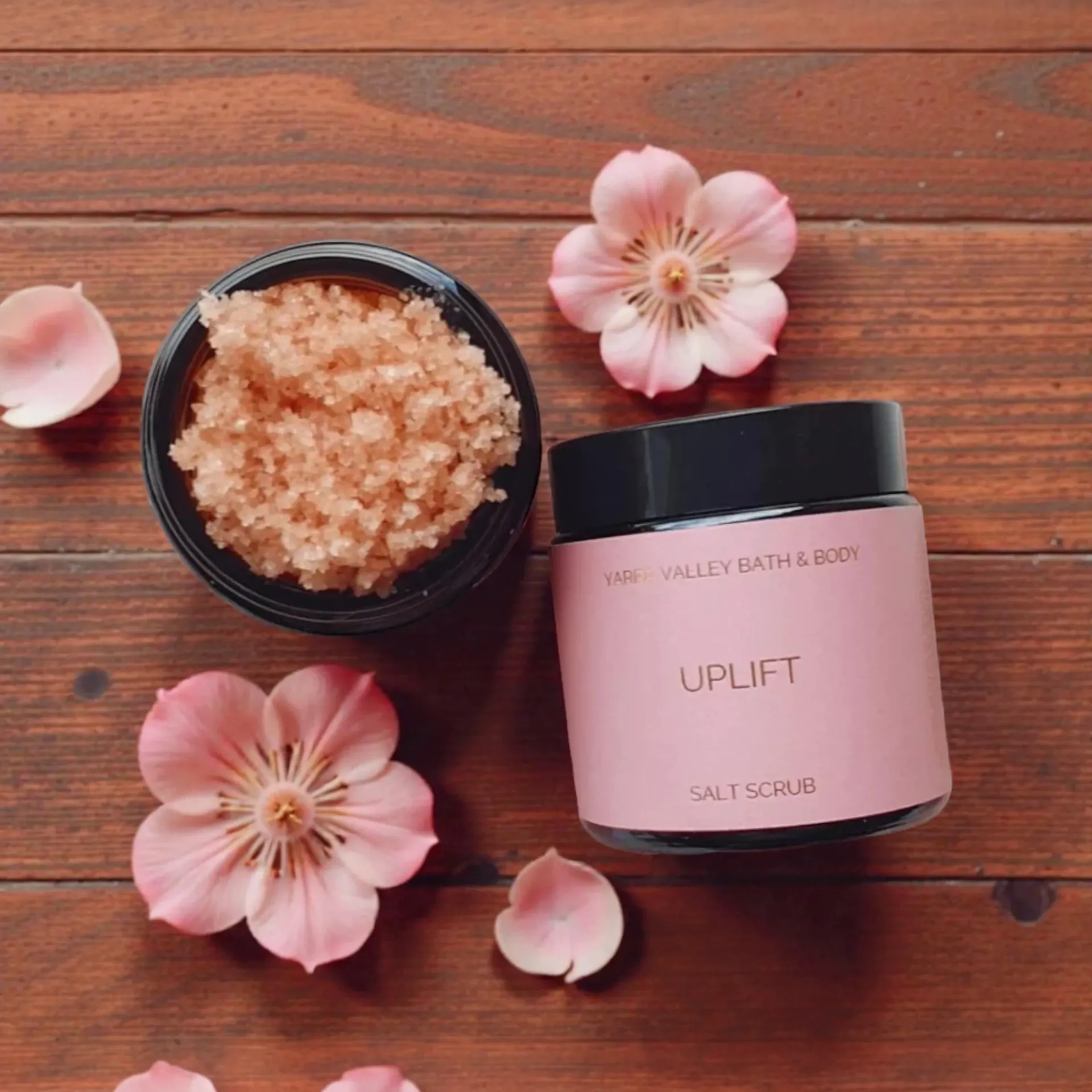 Pink body scrub jar with black lid, bowl of pink crystals, and cherry blossoms on a wooden surface