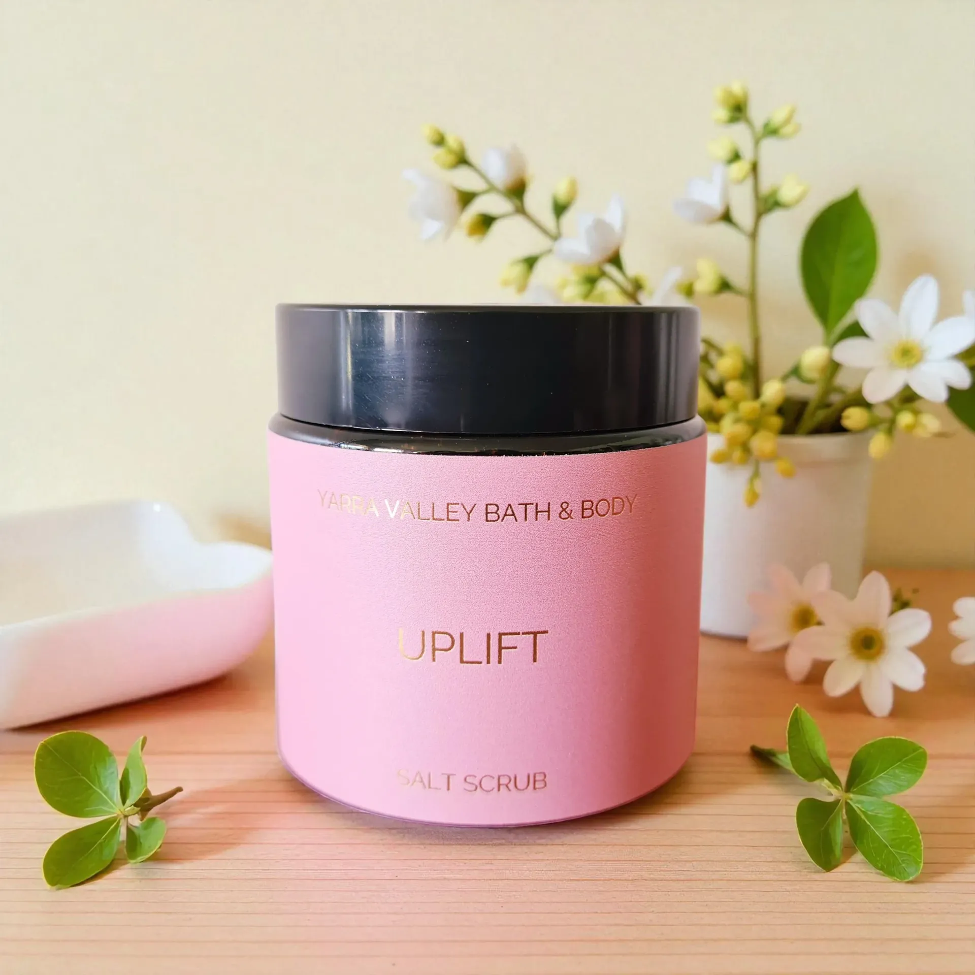 Pink body scrub jar with black lid, bowl of pink crystals, and cherry blossoms on a wooden surface