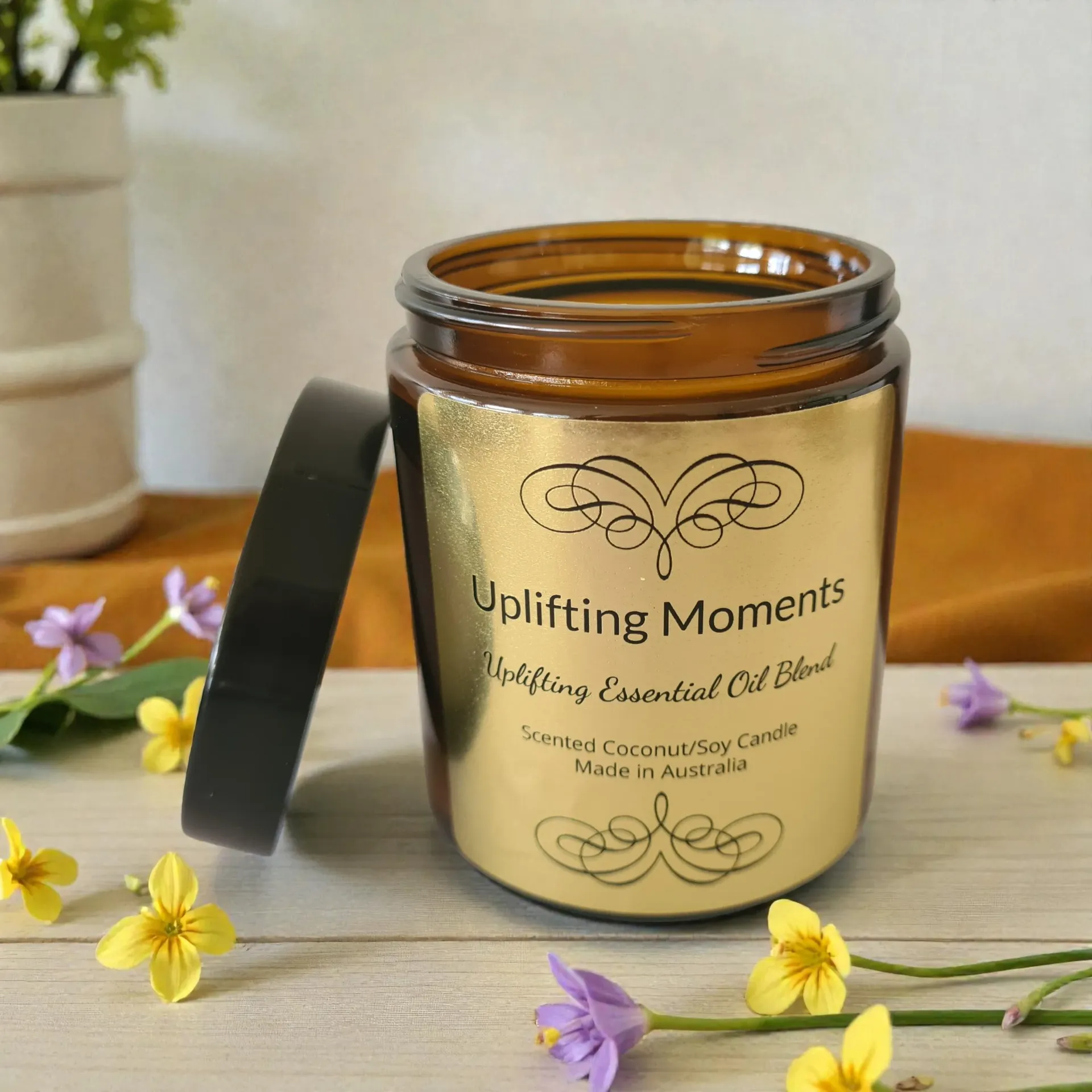 Uplifting Moments scented candle in a gold jar with black lid, set on wood with flowers and green leaves