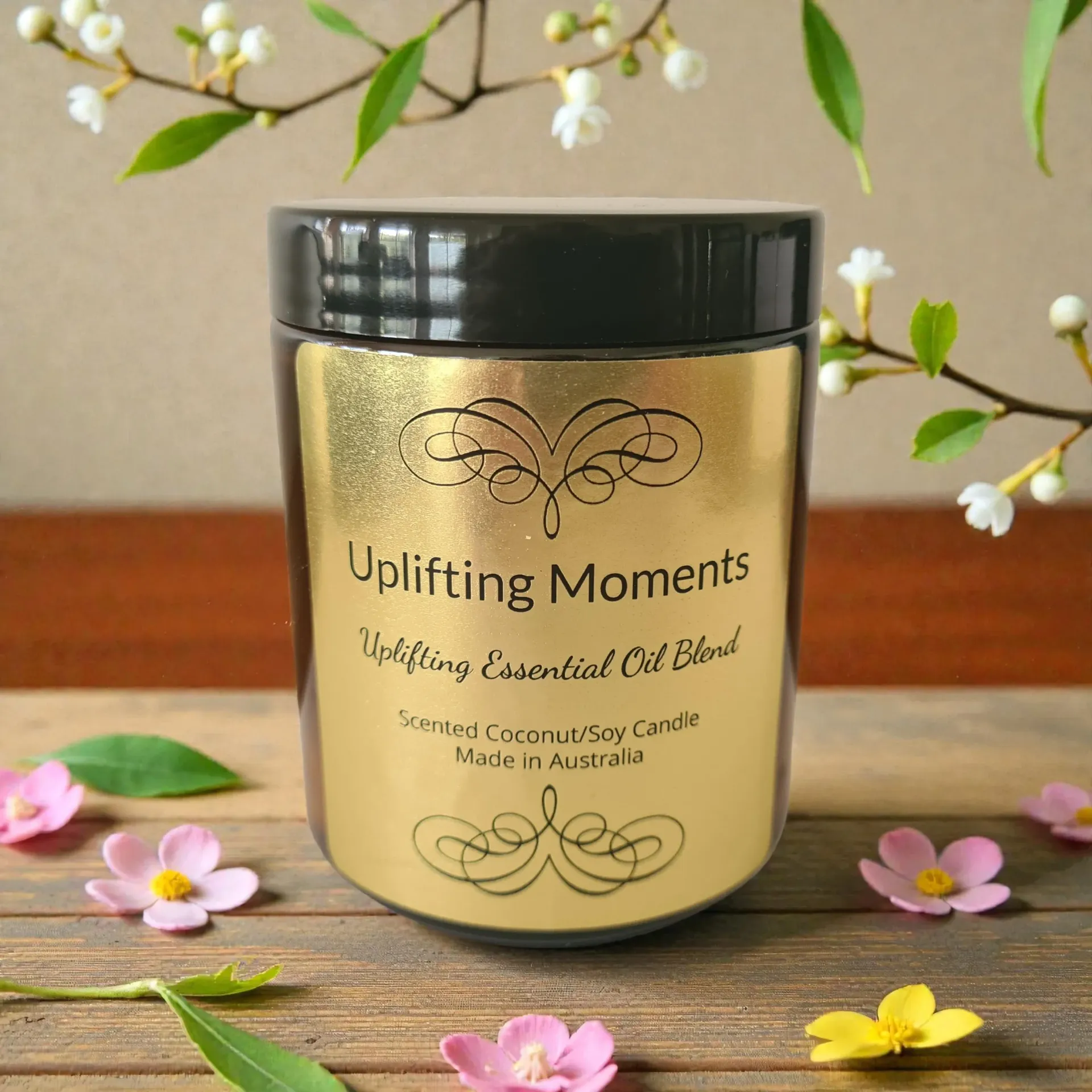 Uplifting Moments scented candle in a gold jar with black lid, set on wood with flowers and green leaves