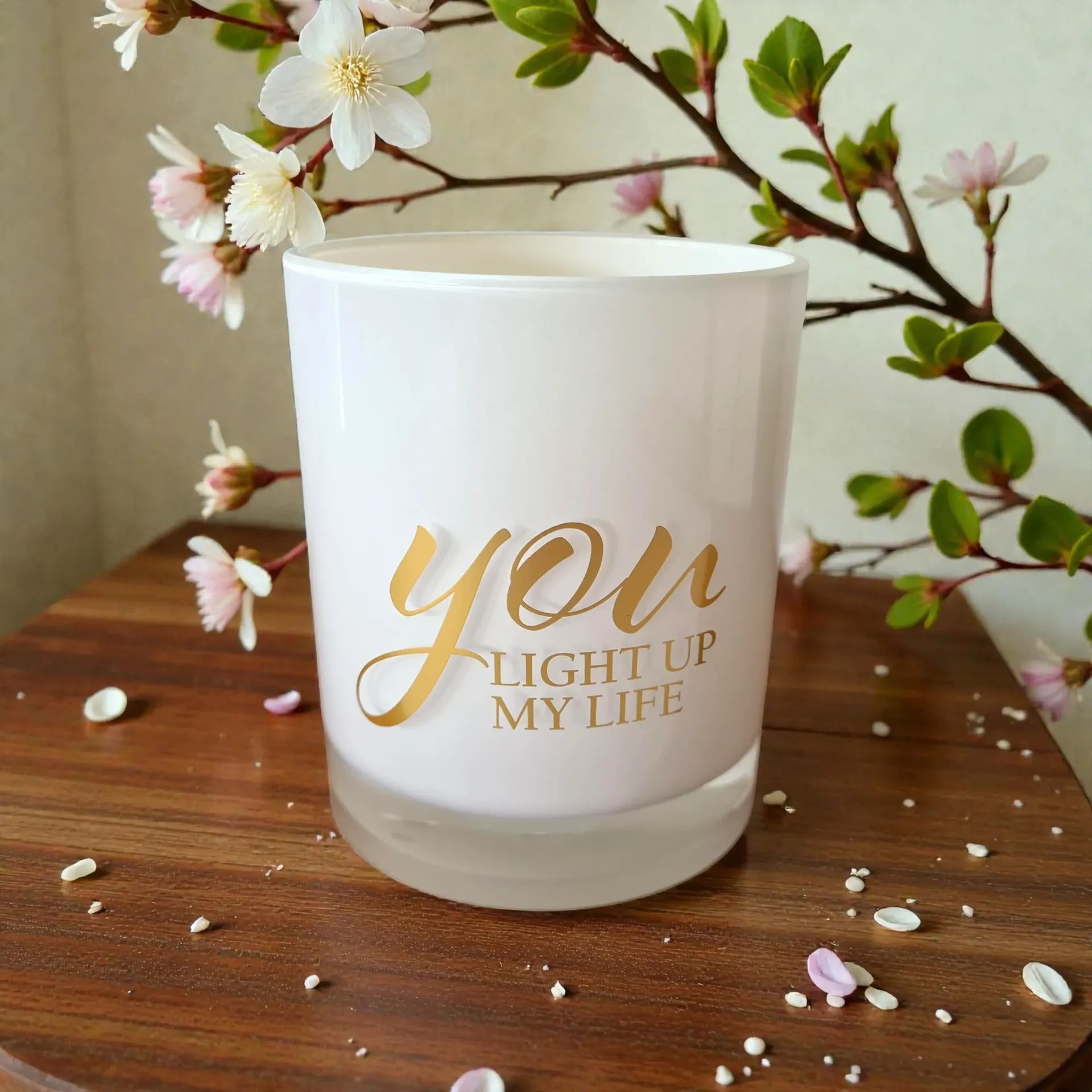 White candle jar with gold “you light up my life” text on a wooden table with blossoms and petals