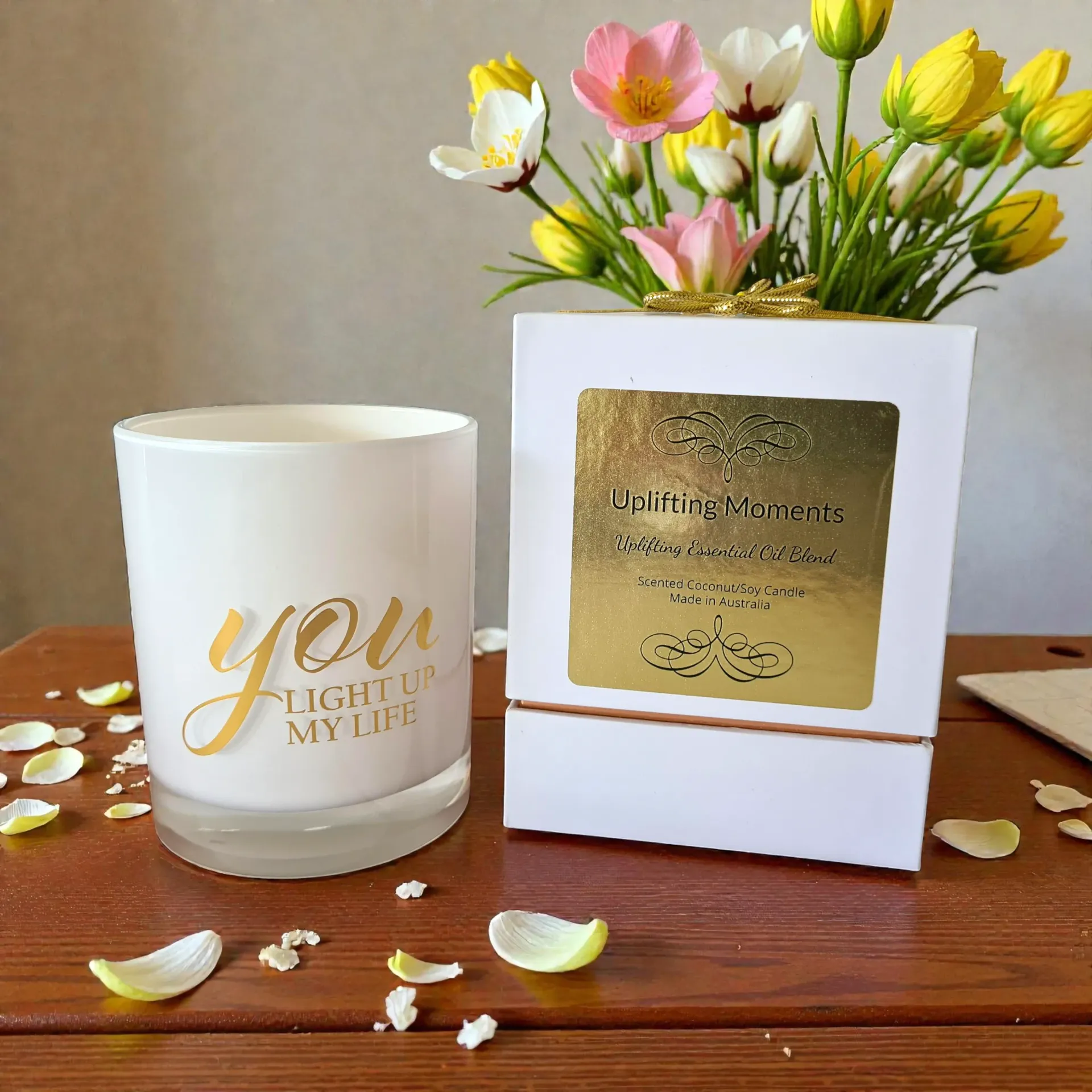 White candle jar with gold “you light up my life” text on a wooden table with blossoms and petals