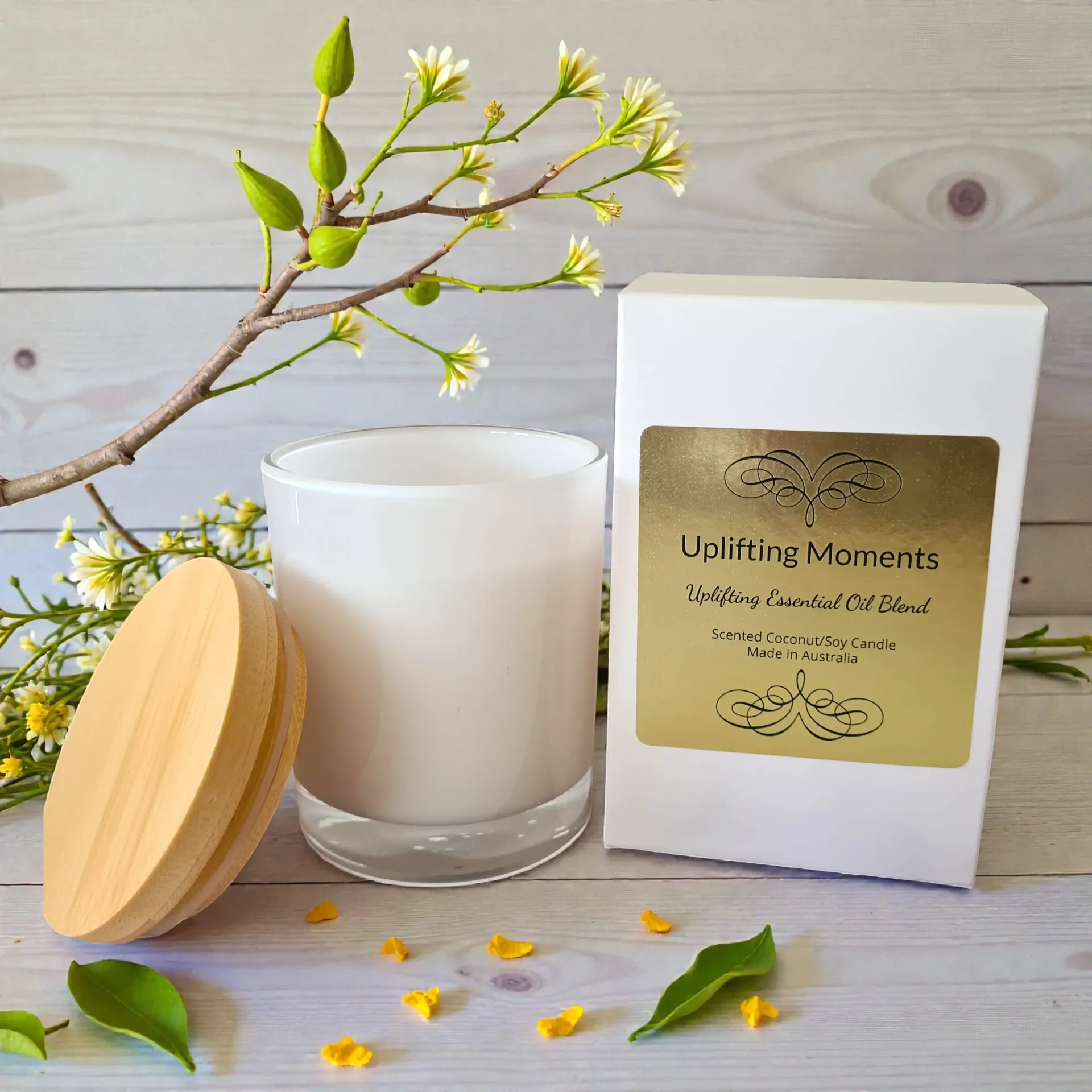 White candle with round wooden lid on a light wood surface, surrounded by small white flowers and green leaves