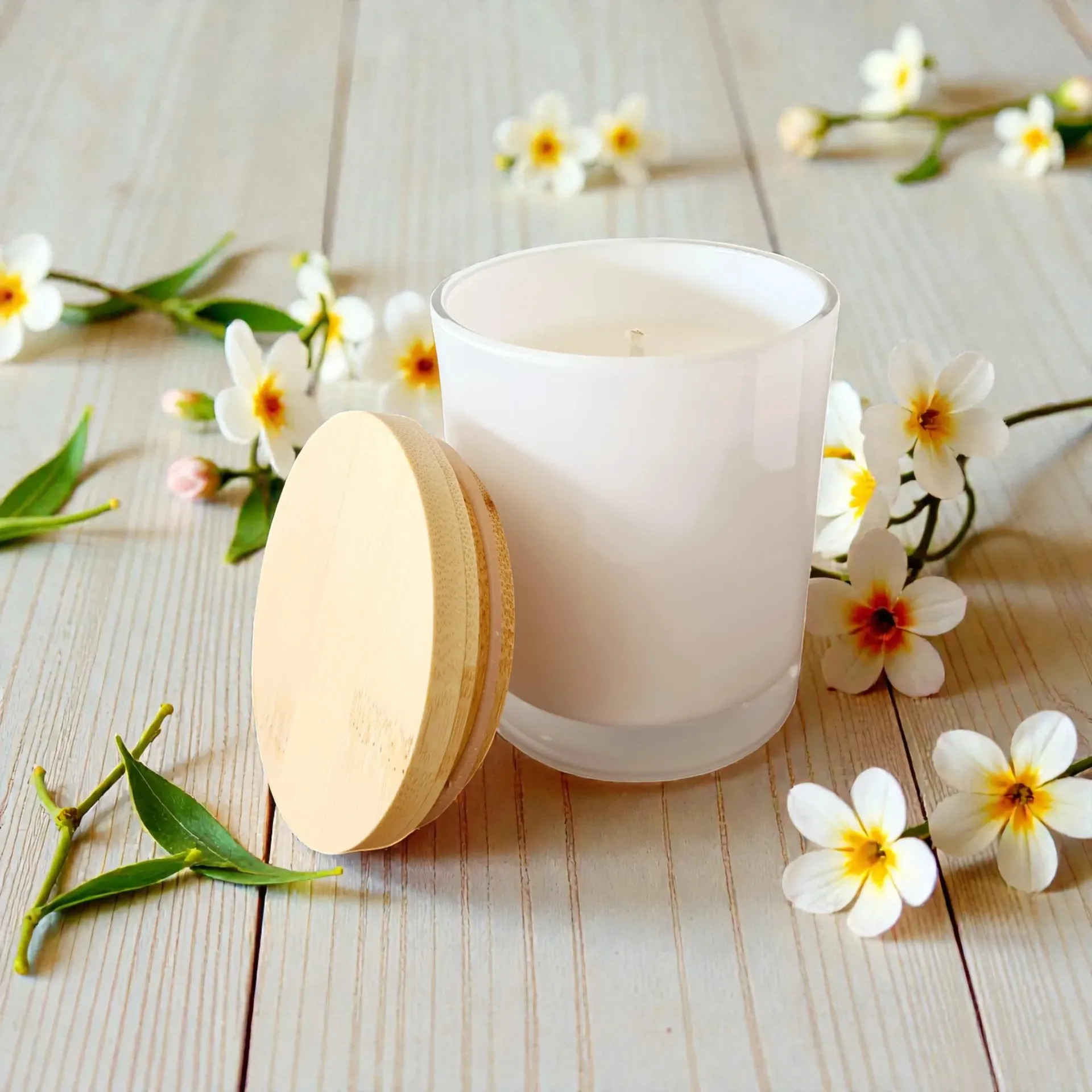 White candle with round wooden lid on a light wood surface, surrounded by small white flowers and green leaves