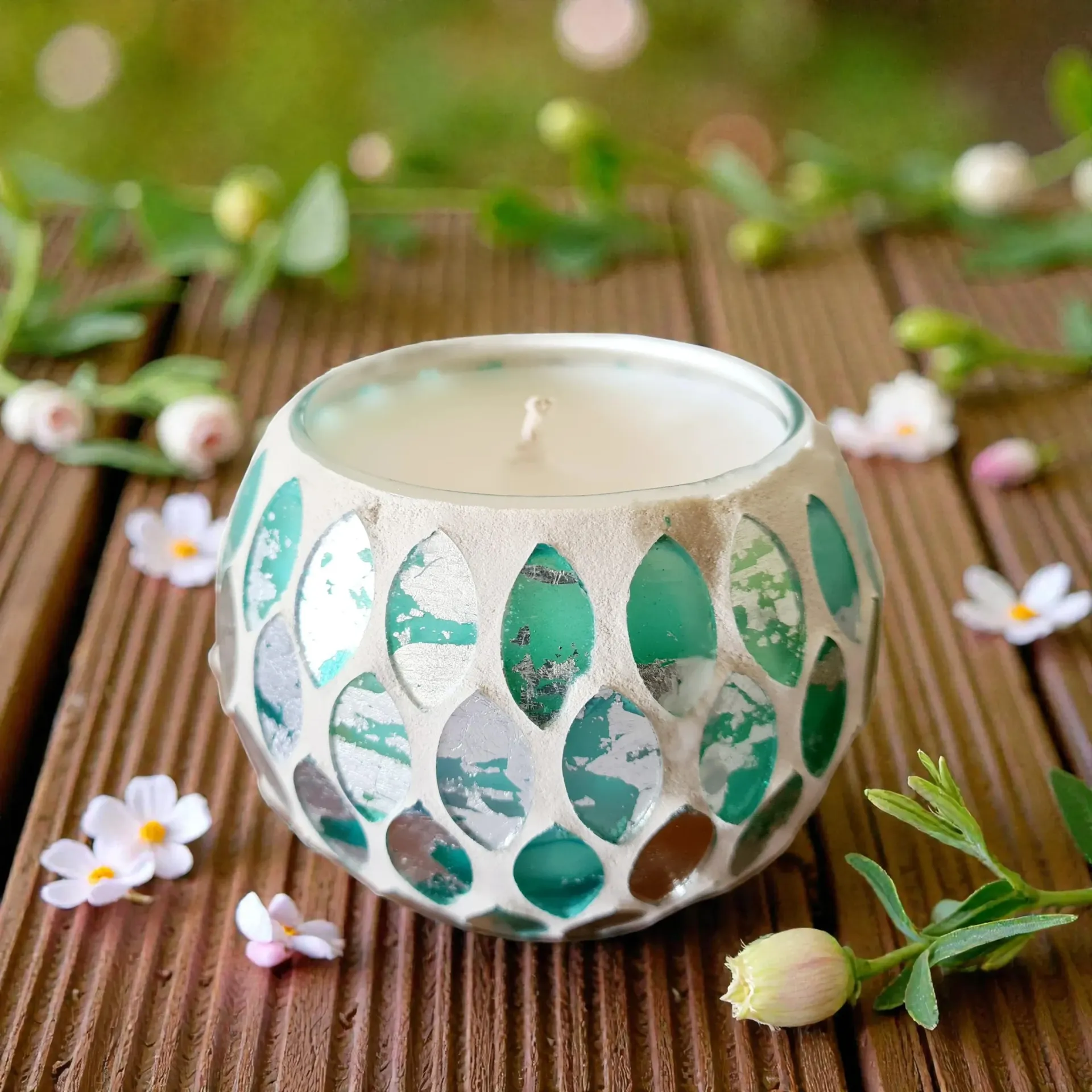 White candle in a teal mosaic glass holder on a wooden surface, surrounded by small white flowers and greenery