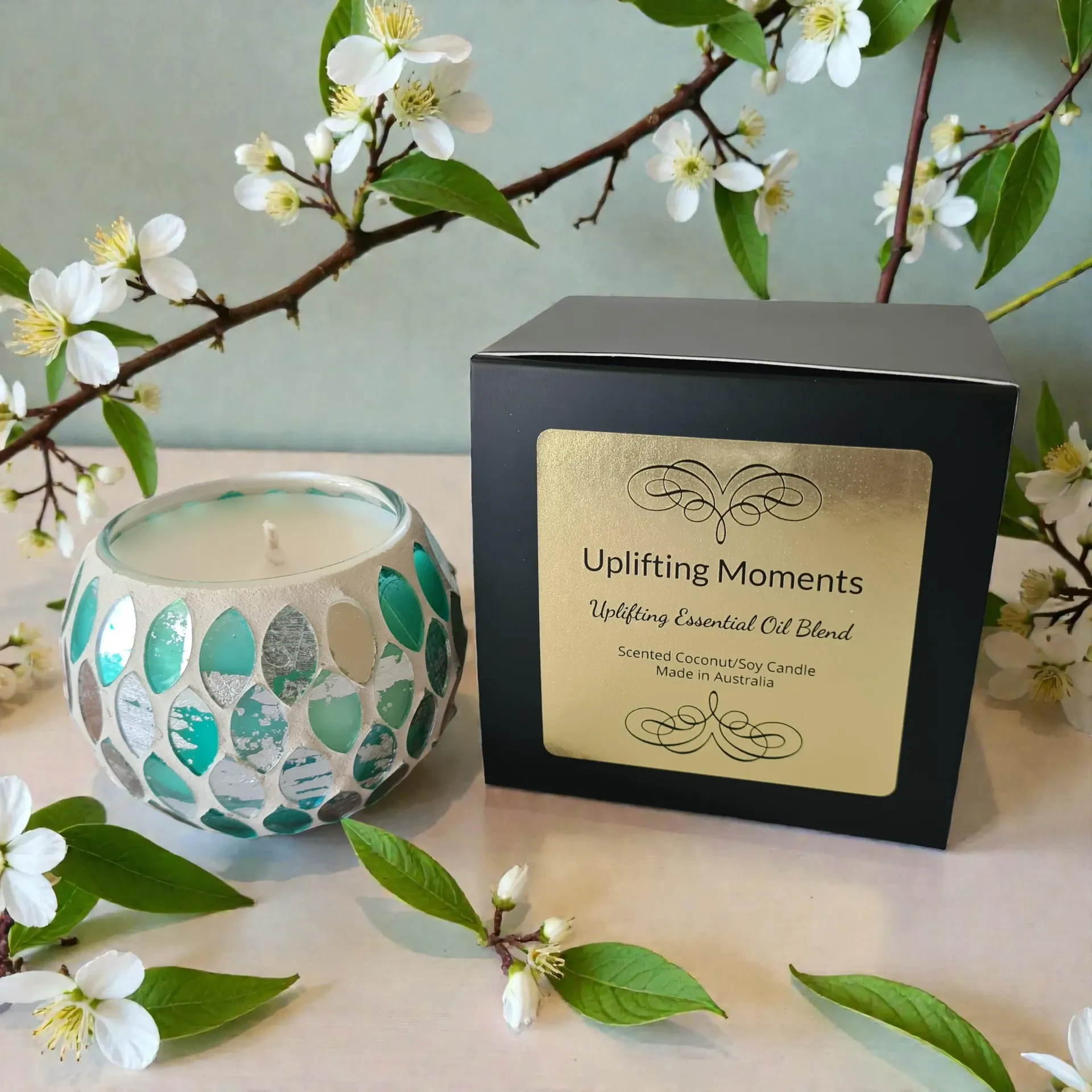 White candle in a teal mosaic glass holder on a wooden surface, surrounded by small white flowers and greenery