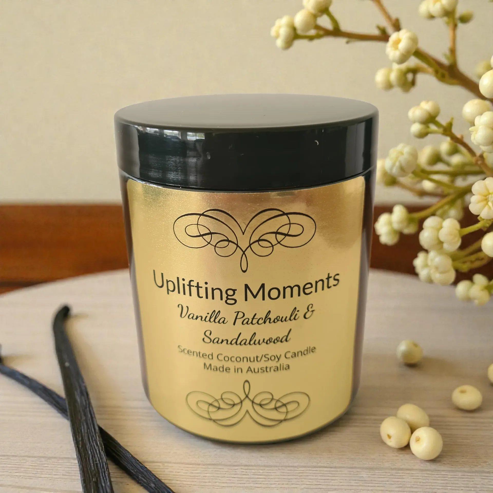 Amber candle jar labeled “Uplifting Moments” with a black lid on a wooden surface, beside white flowers.
