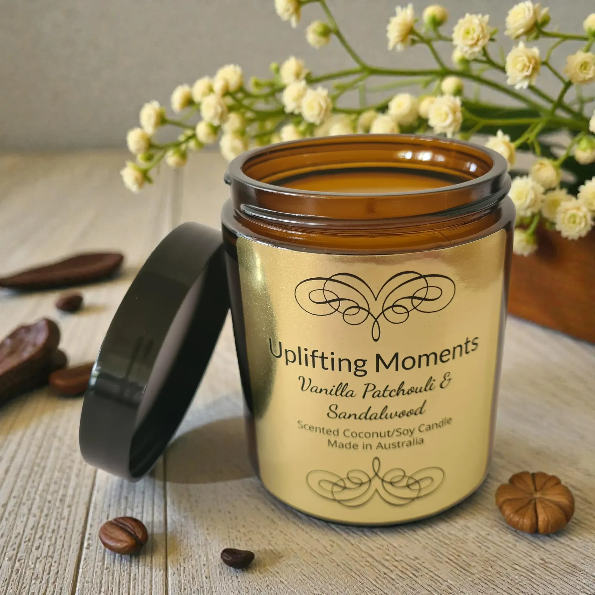 Amber candle jar labeled “Uplifting Moments” with a black lid on a wooden surface, beside white flowers.