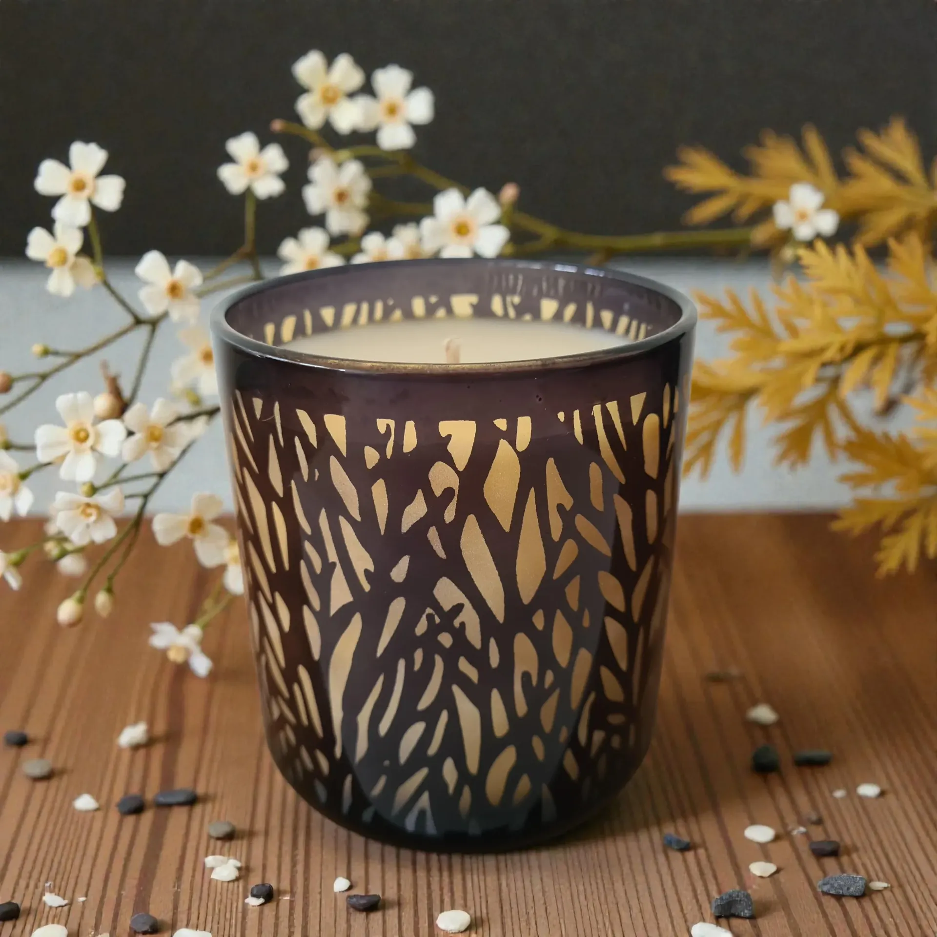 Decorative dark glass candle holder on a wooden table with white flowers and scattered pebbles