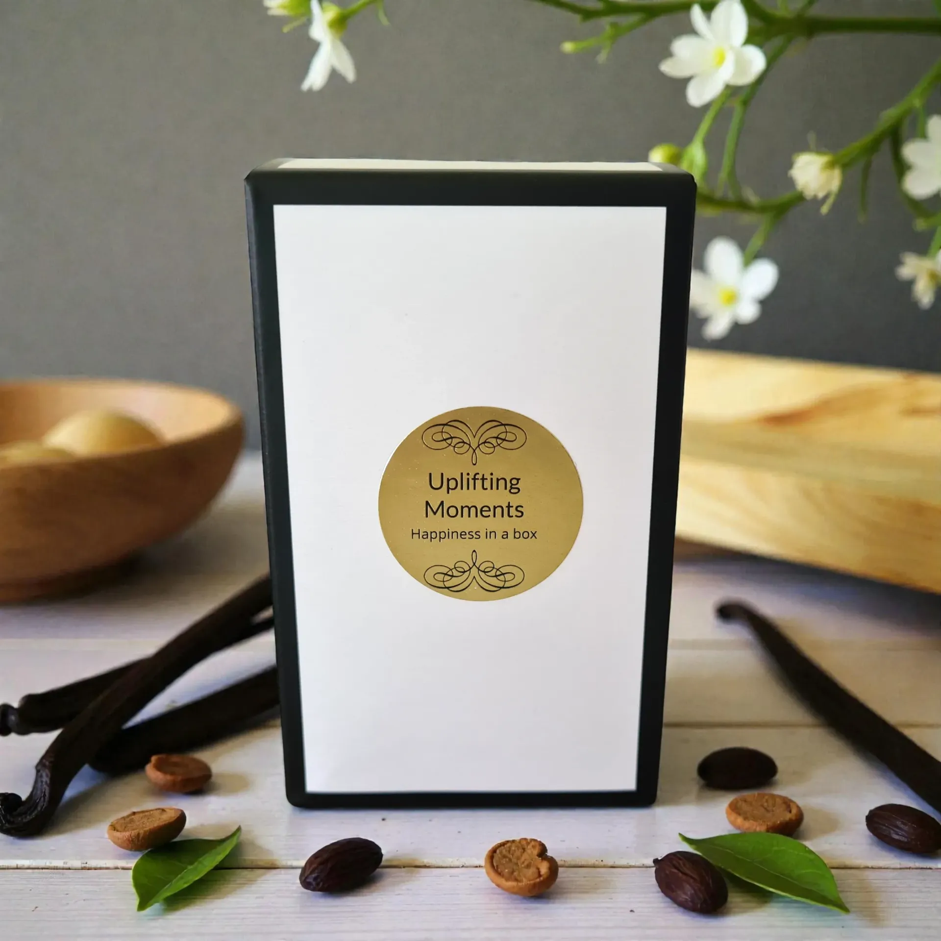 Frosted candle with “Uplifting Moments” label, surrounded by vanilla beans, coffee beans, and flowers on a tabletop