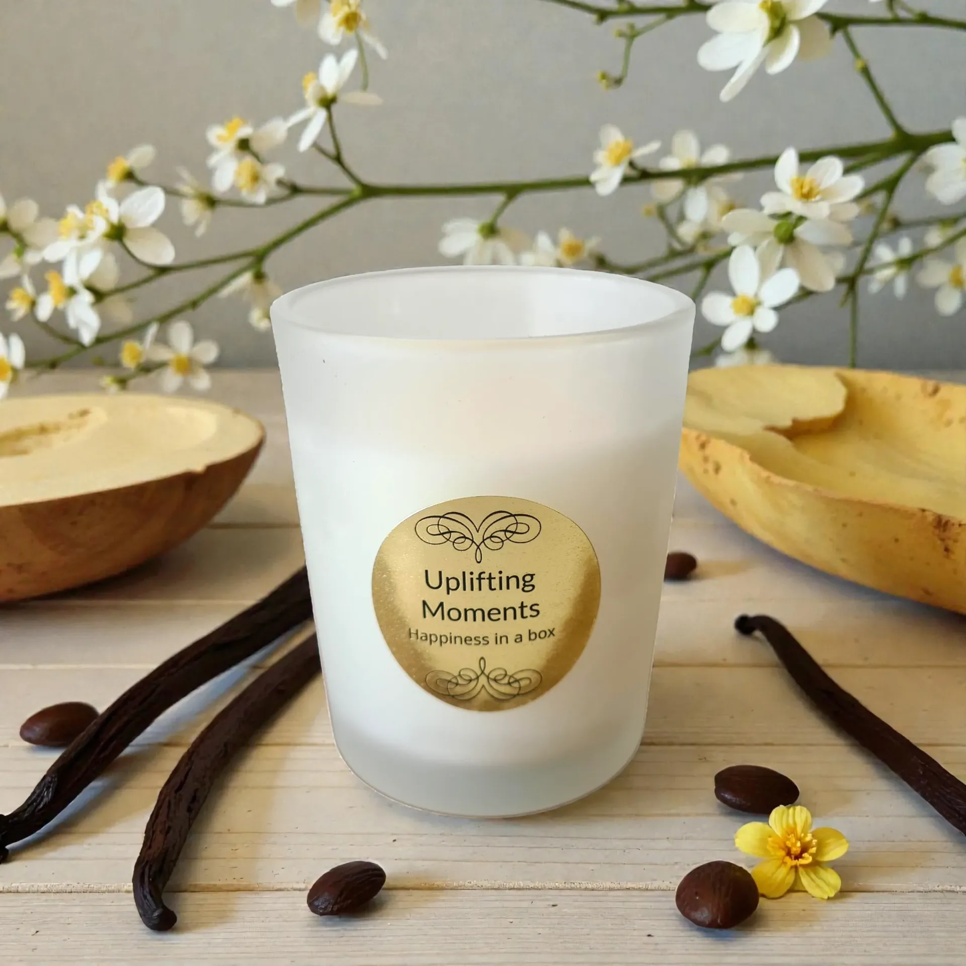 Frosted candle with “Uplifting Moments” label, surrounded by vanilla beans, coffee beans, and flowers on a tabletop