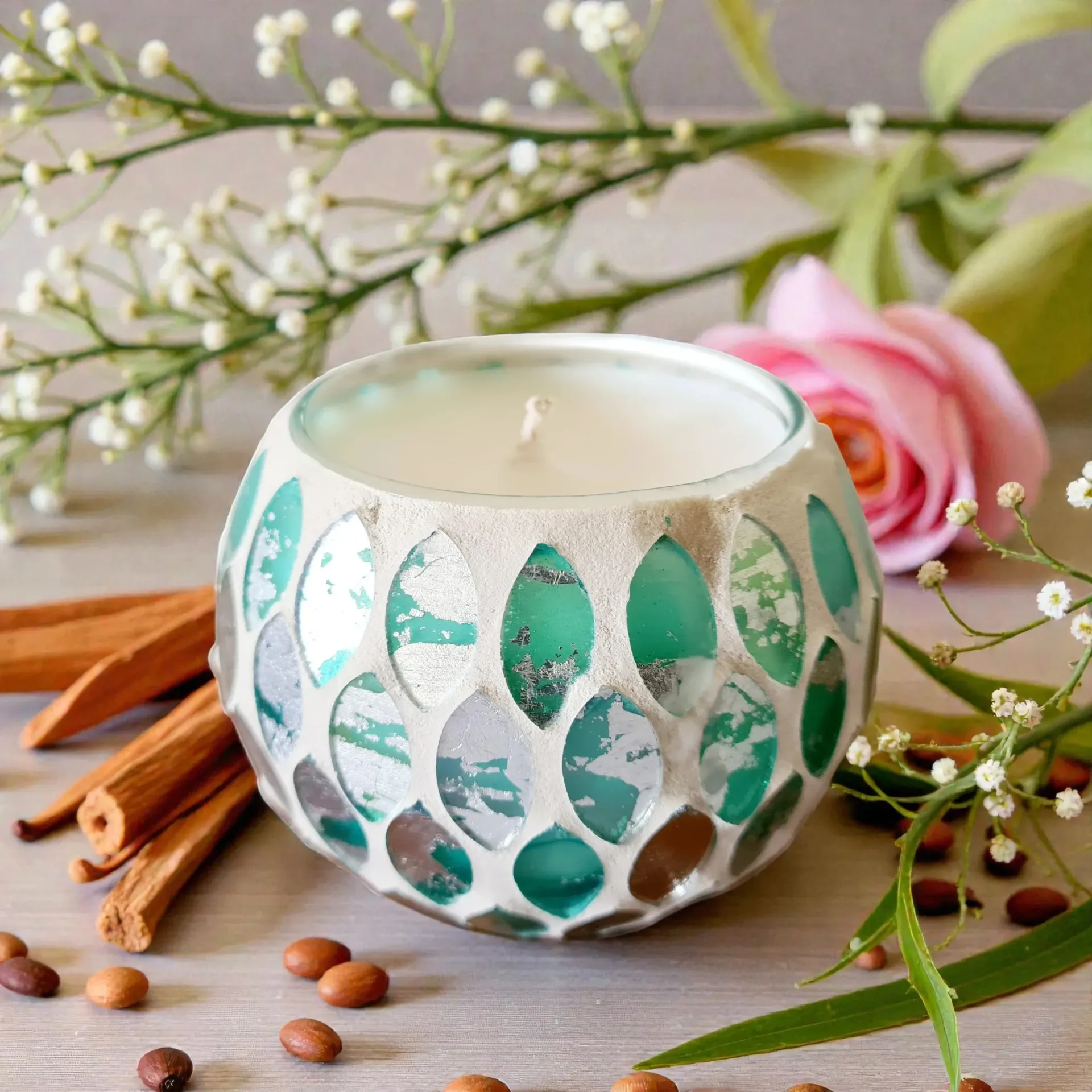Decorative teal mosaic candle holder with white candle, cinnamon sticks, and a pink rose on a tabletop