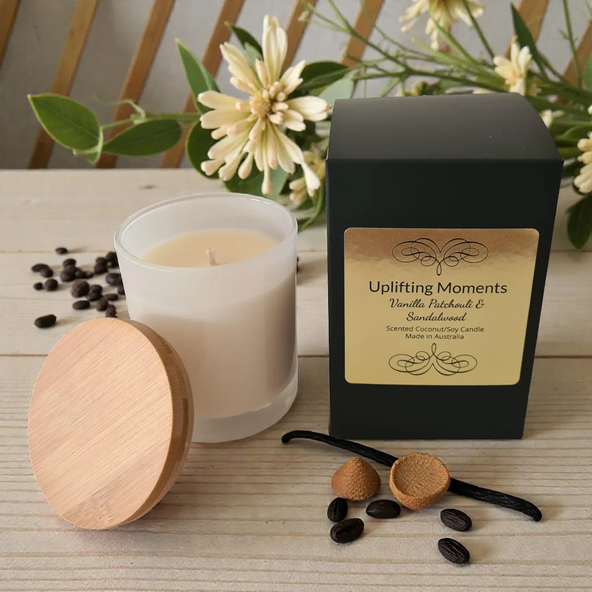 White candle with wooden lid beside black aromatherapy box, scattered coffee beans, and white flowers on a table