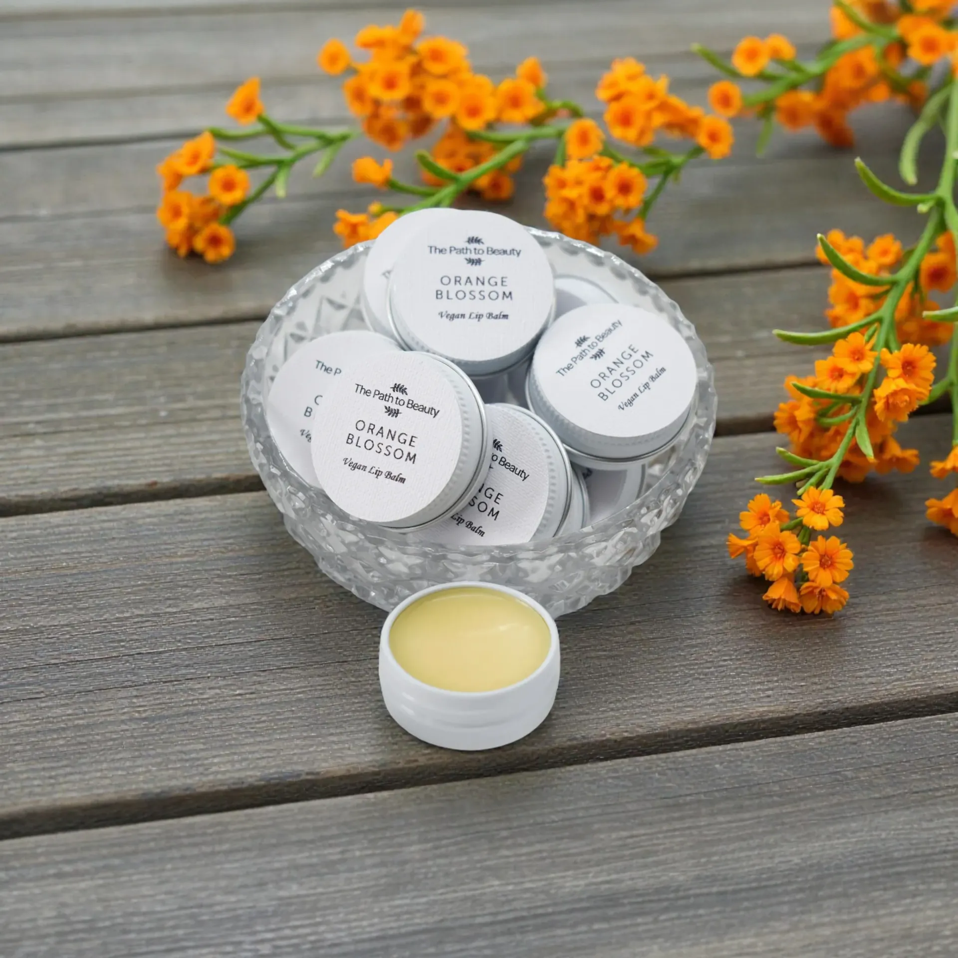 Glass bowl of white vegan lip balm tins with orange flowers on a gray wooden table