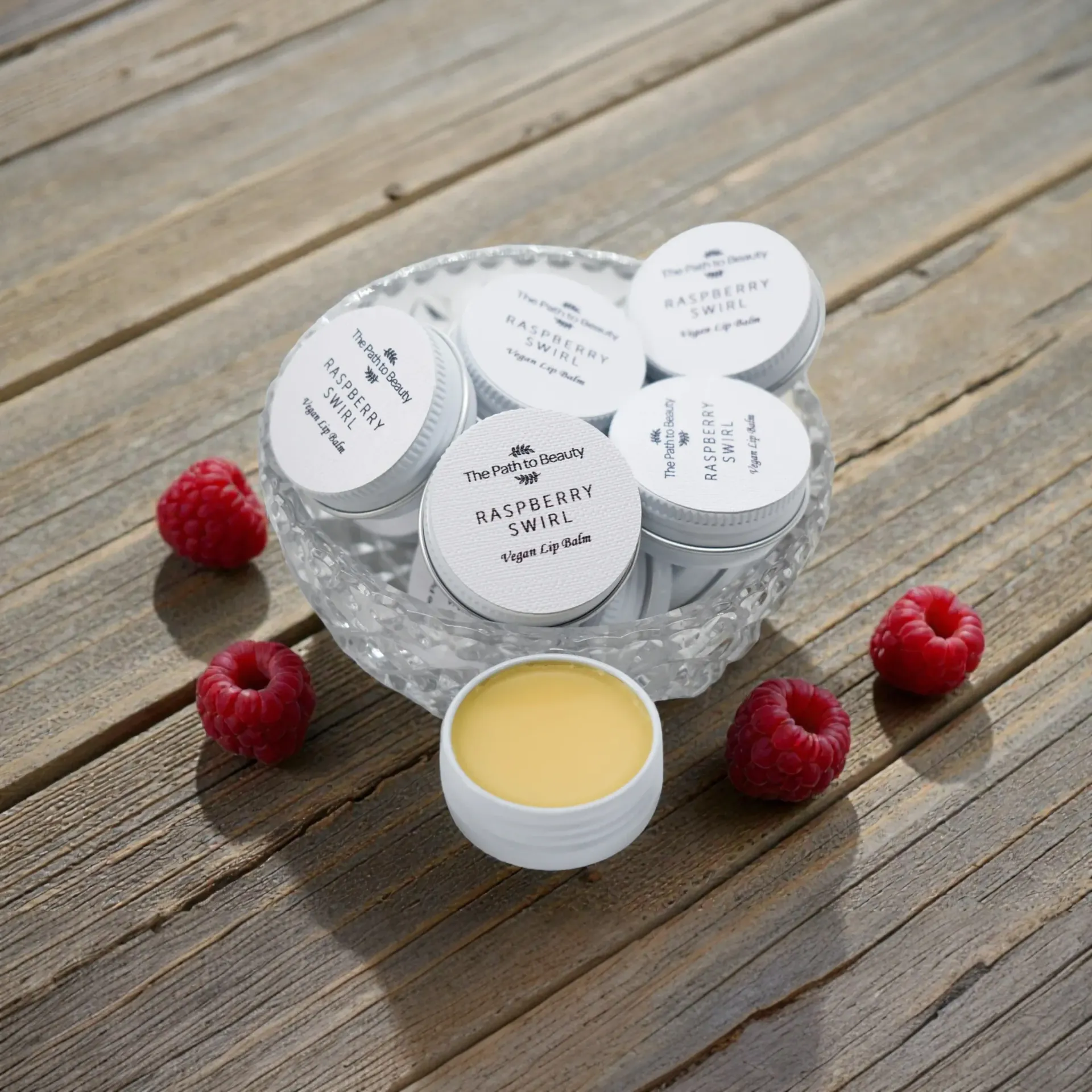 Small white vegan lip balm jars in a glass bowl on a wooden table, with raspberries around them