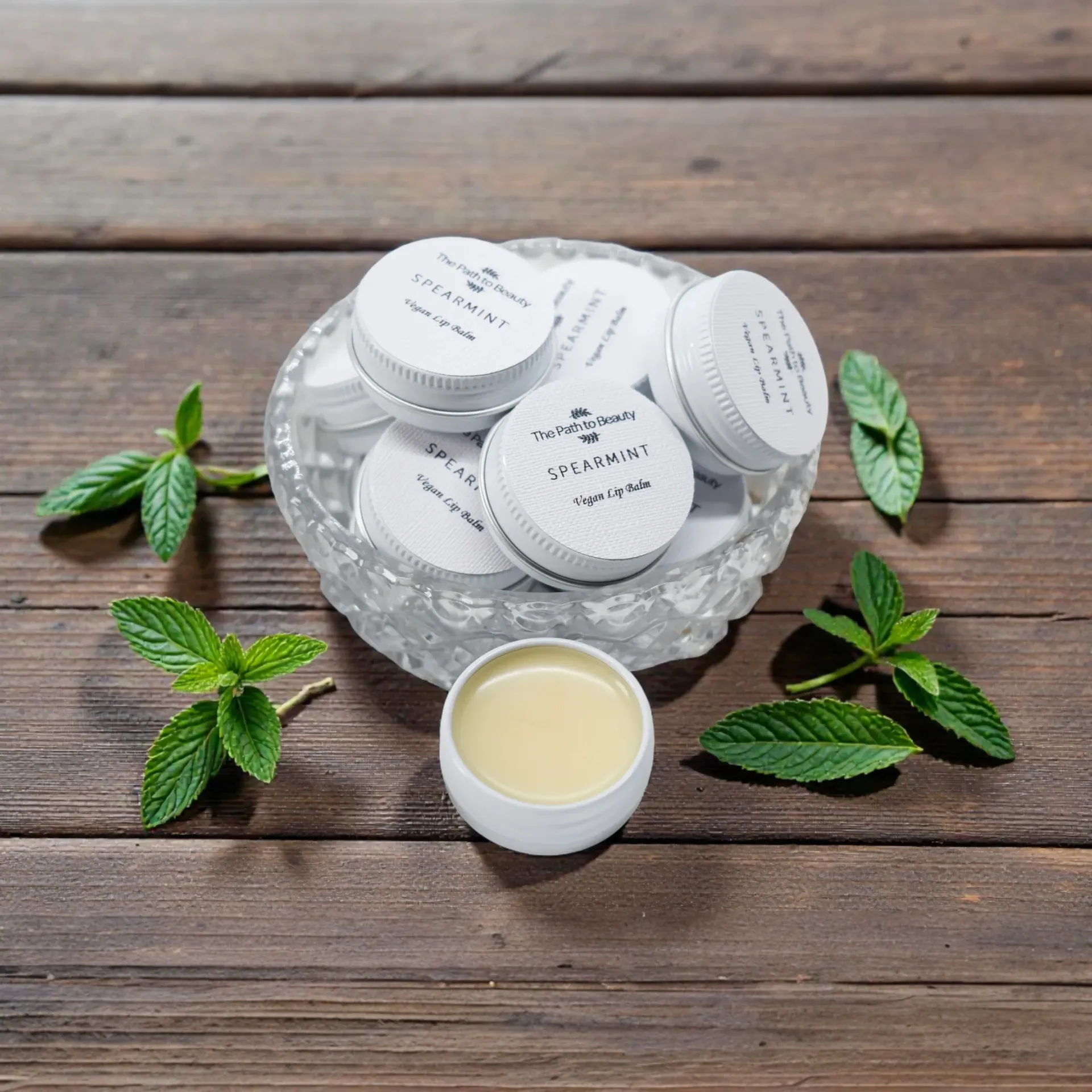 White cosmetic vegan lip balm jars in a glass bowl on a wooden table, surrounded by green mint leaves
