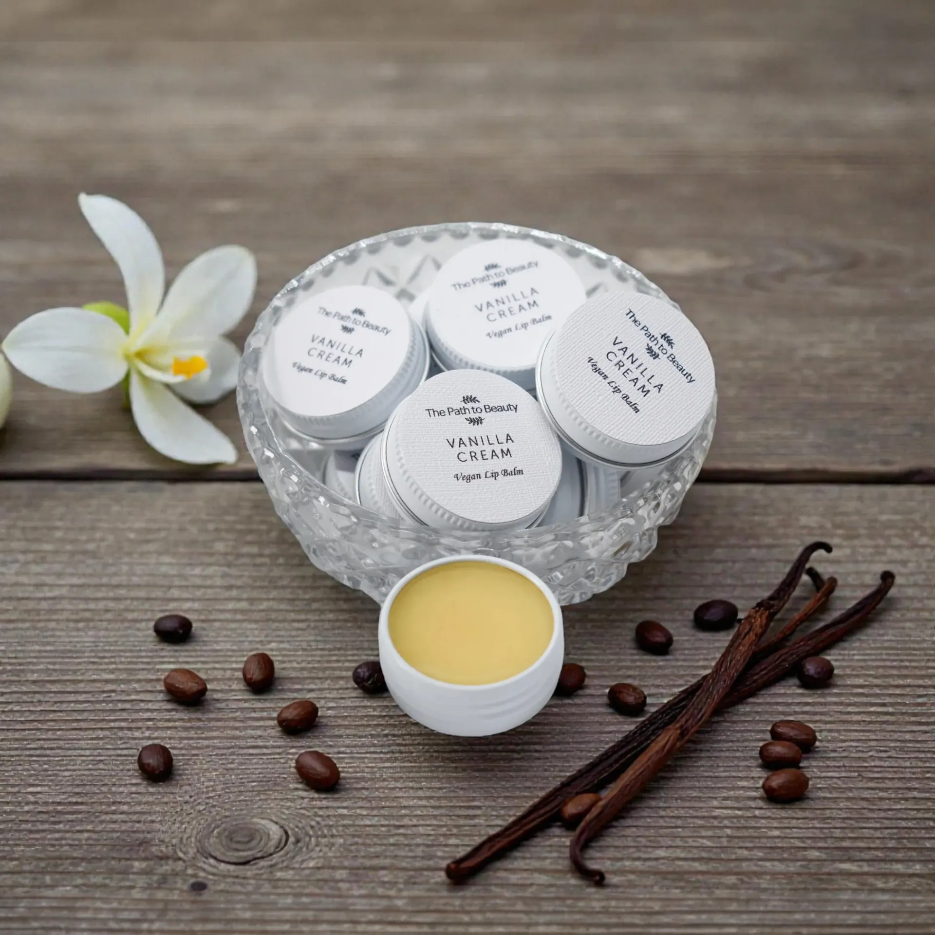 Open basket of white vegan lip balm jars with a yellow cream pot, coffee beans, vanilla pods, and a white flower on wood.
