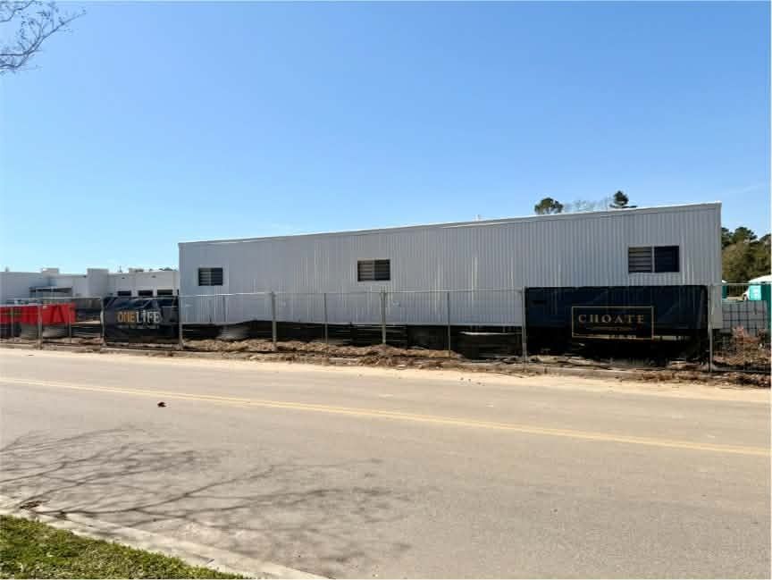 White industrial building beside a road, with construction equipment and a clear blue sky.