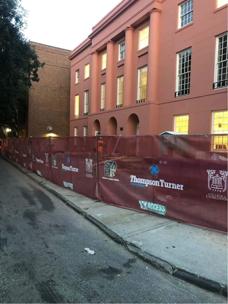 Red brick building behind a maroon construction fence along a sidewalk at dusk